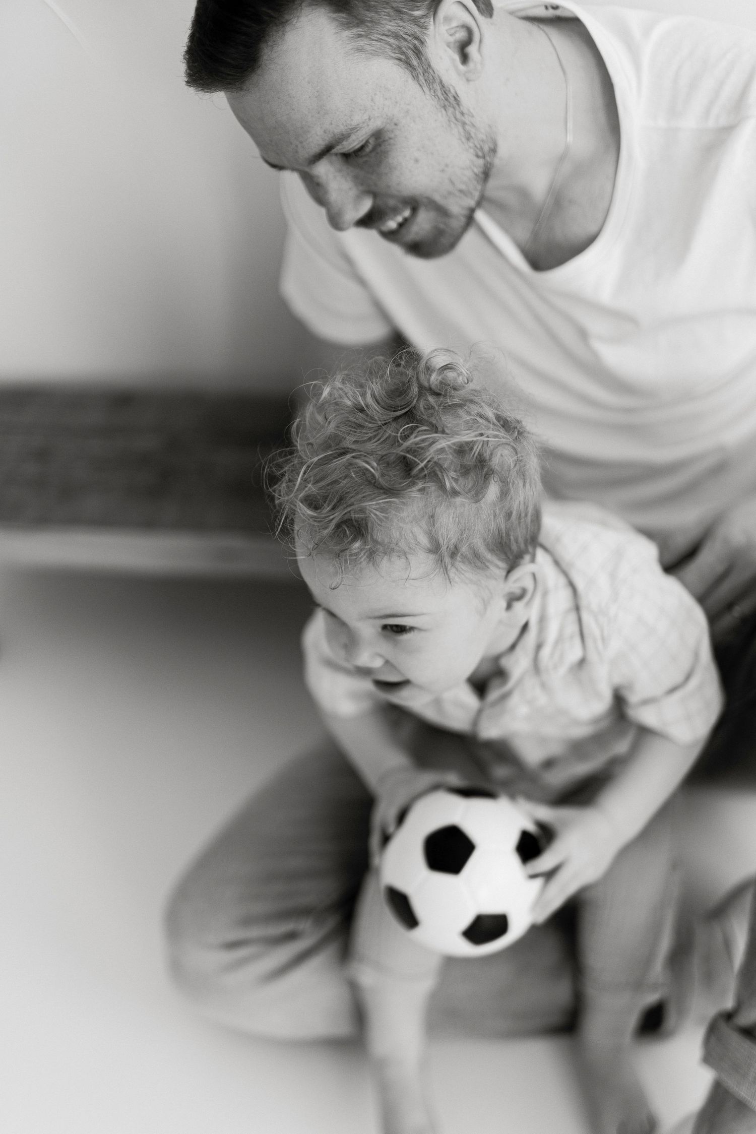 Black and white photo of a father sitting on floor with toddler holding a soccer ball.