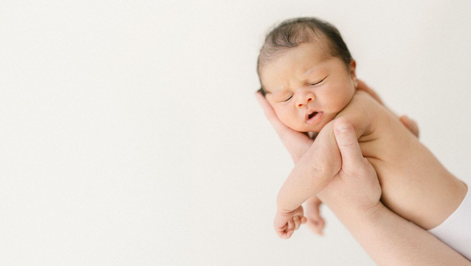 A newborn baby with dark hair sleeping peacefully while being gently cradled in adult hands against white background.