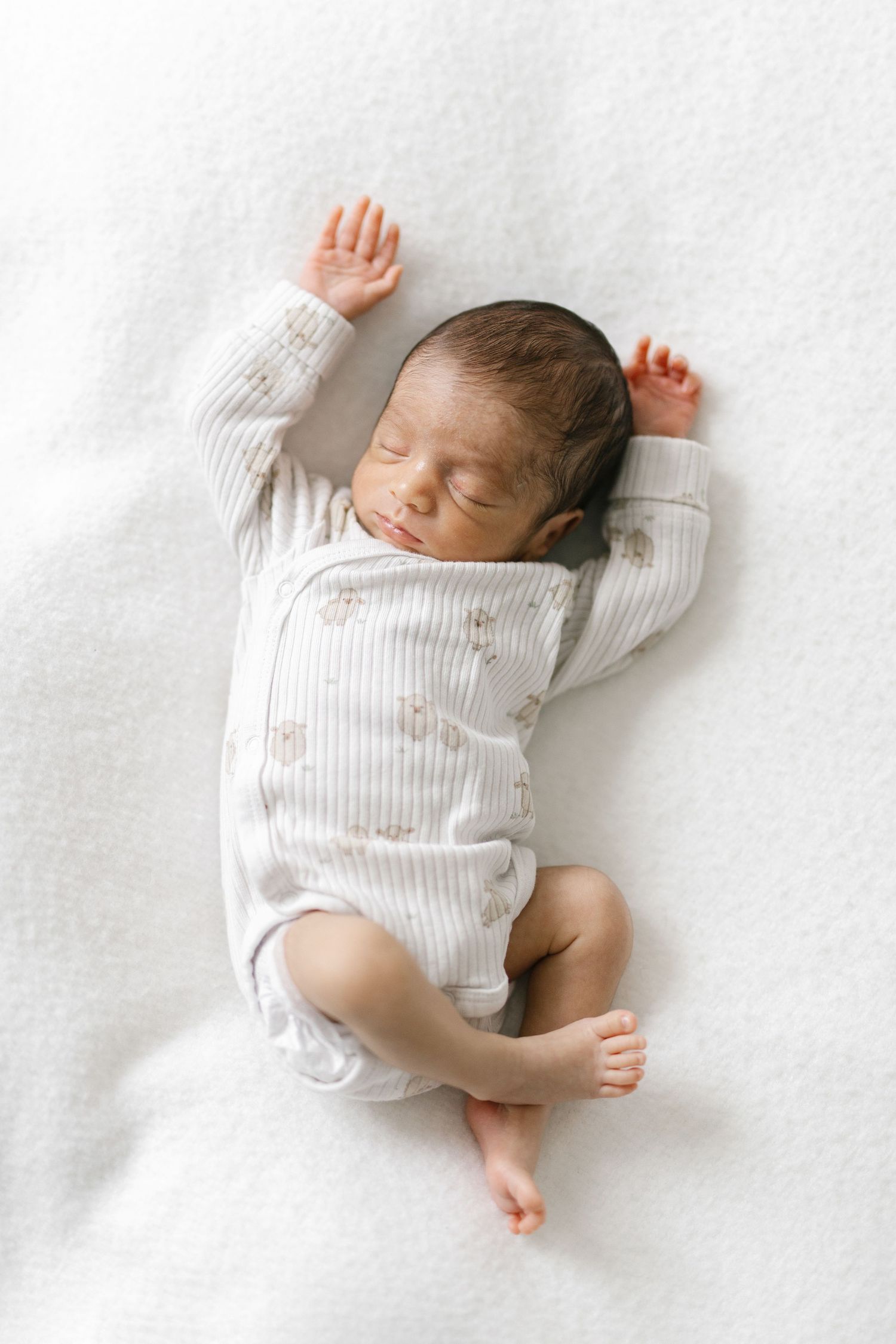 Peaceful newborn baby sleeping on white blanket wearing striped onesie with tiny floral print, arms raised overhead.
