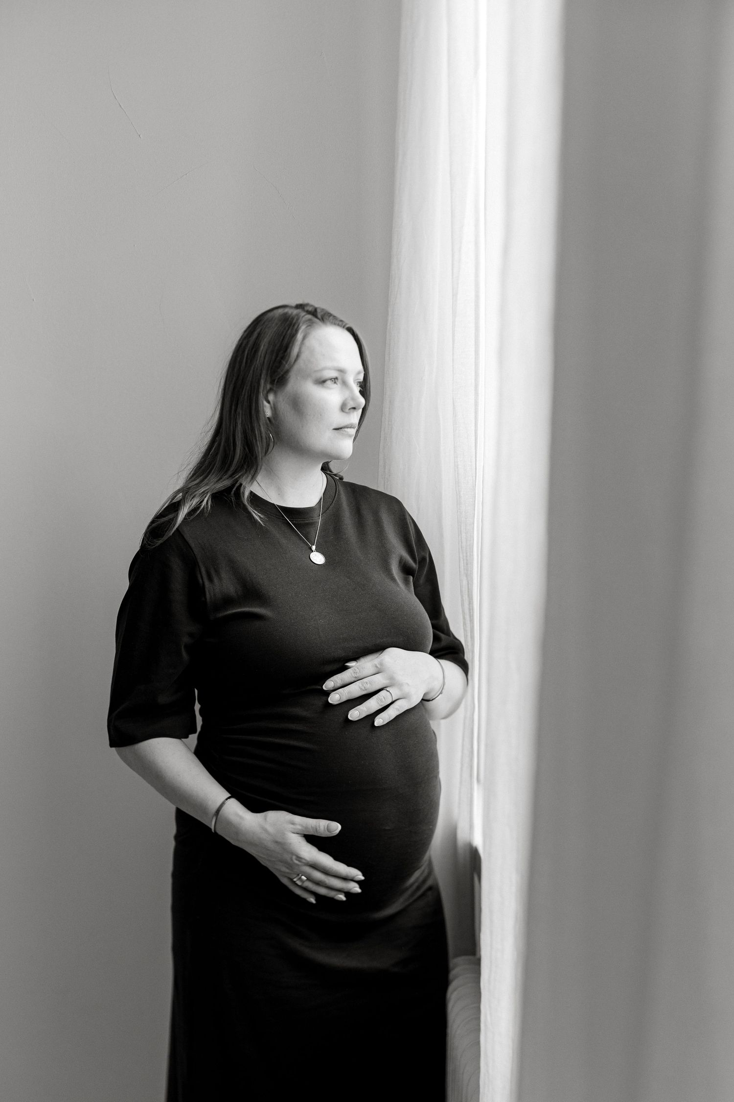 Black and white photo of a pregnant woman in a black dress cradling her baby bump while standing near a window.
