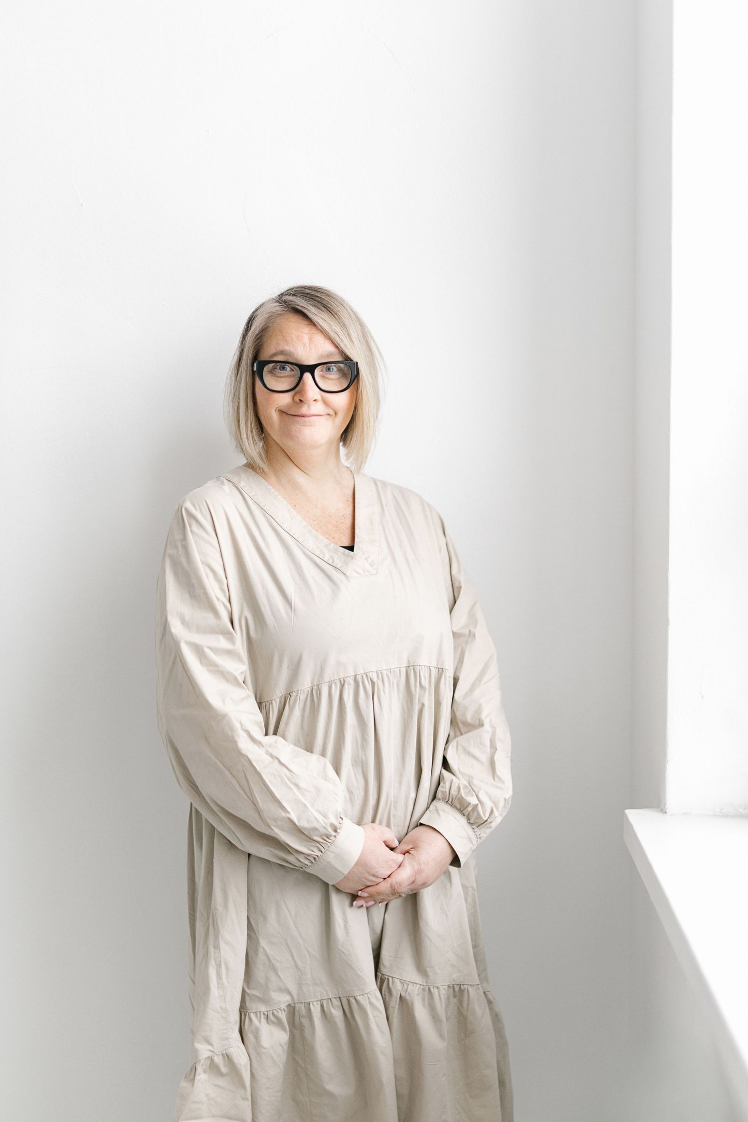 Person wearing glasses and a beige tiered maxi dress stands against white wall in minimalist setting.