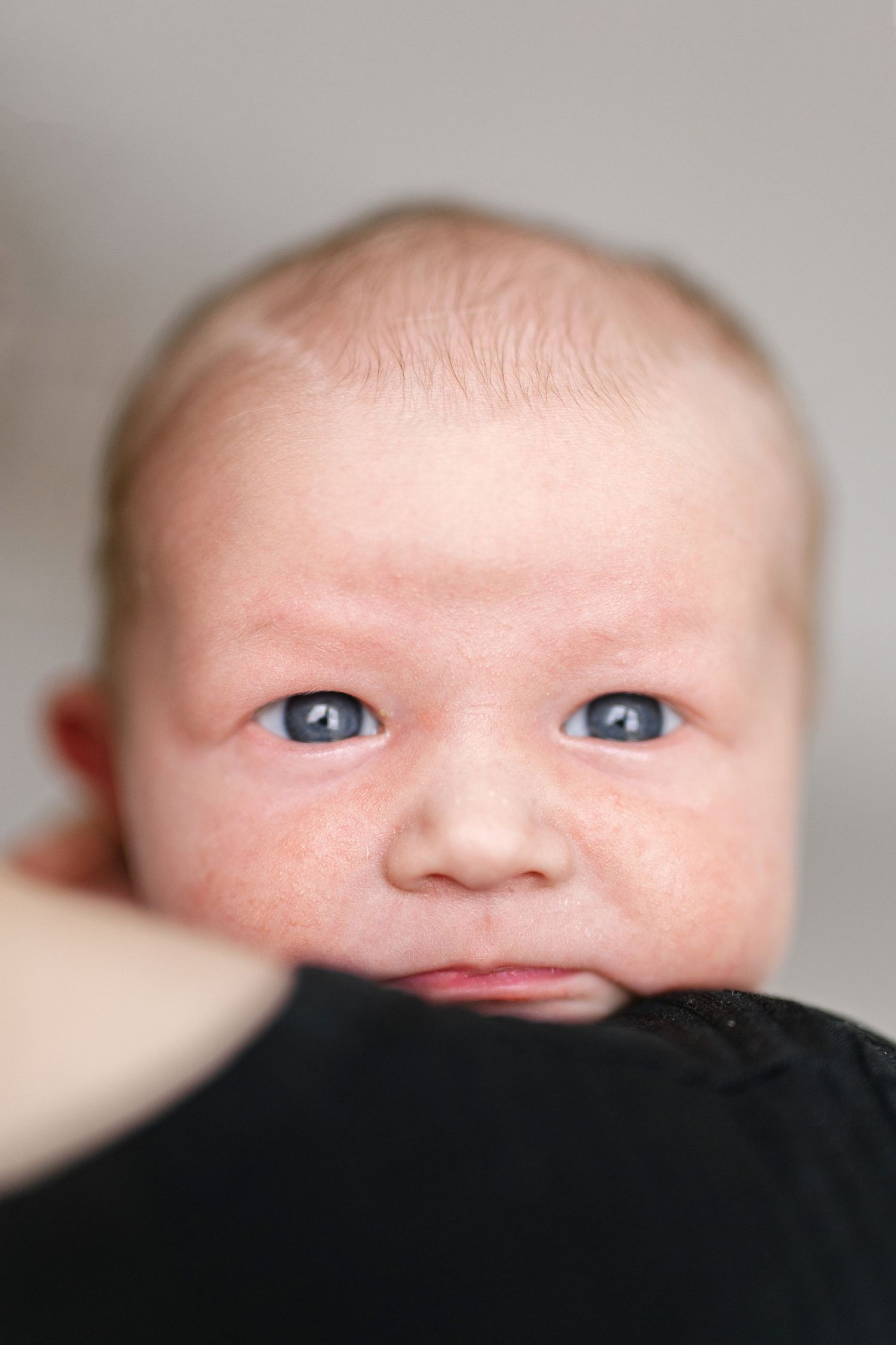 Close-up portrait of a newborn baby with blue eyes resting on a parent's shoulder in soft light.