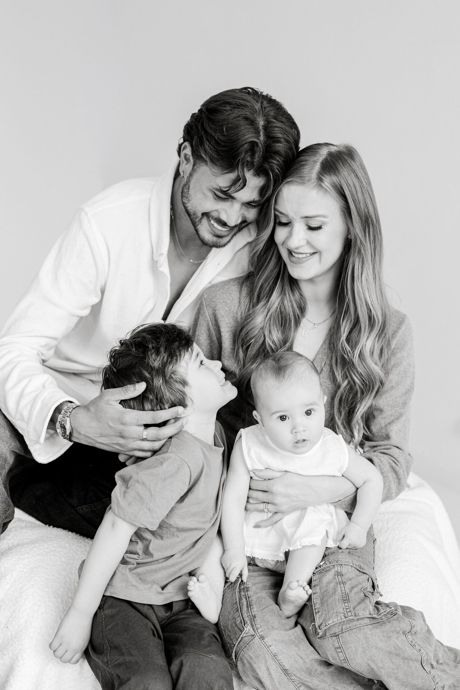 Black and white family portrait of parents with young child and baby sitting together, smiling warmly in studio setting.
