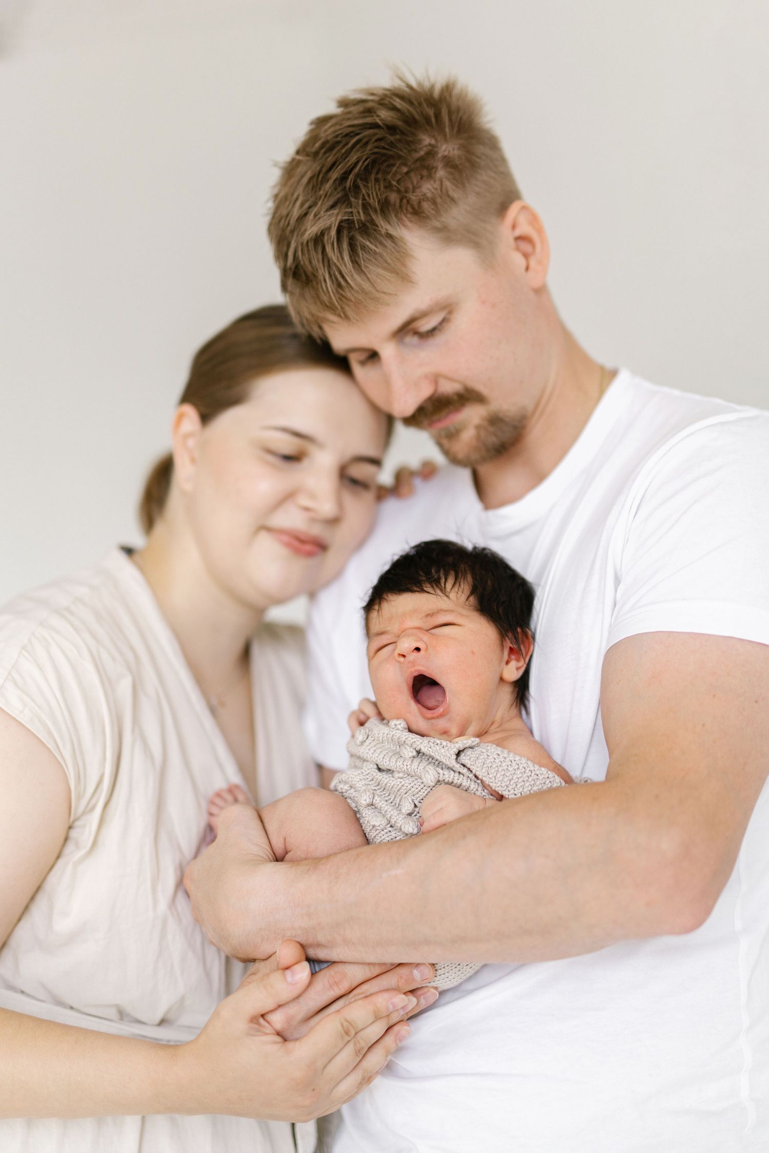 New parents tenderly hold their yawning newborn baby, sharing a precious family moment in soft, neutral tones.