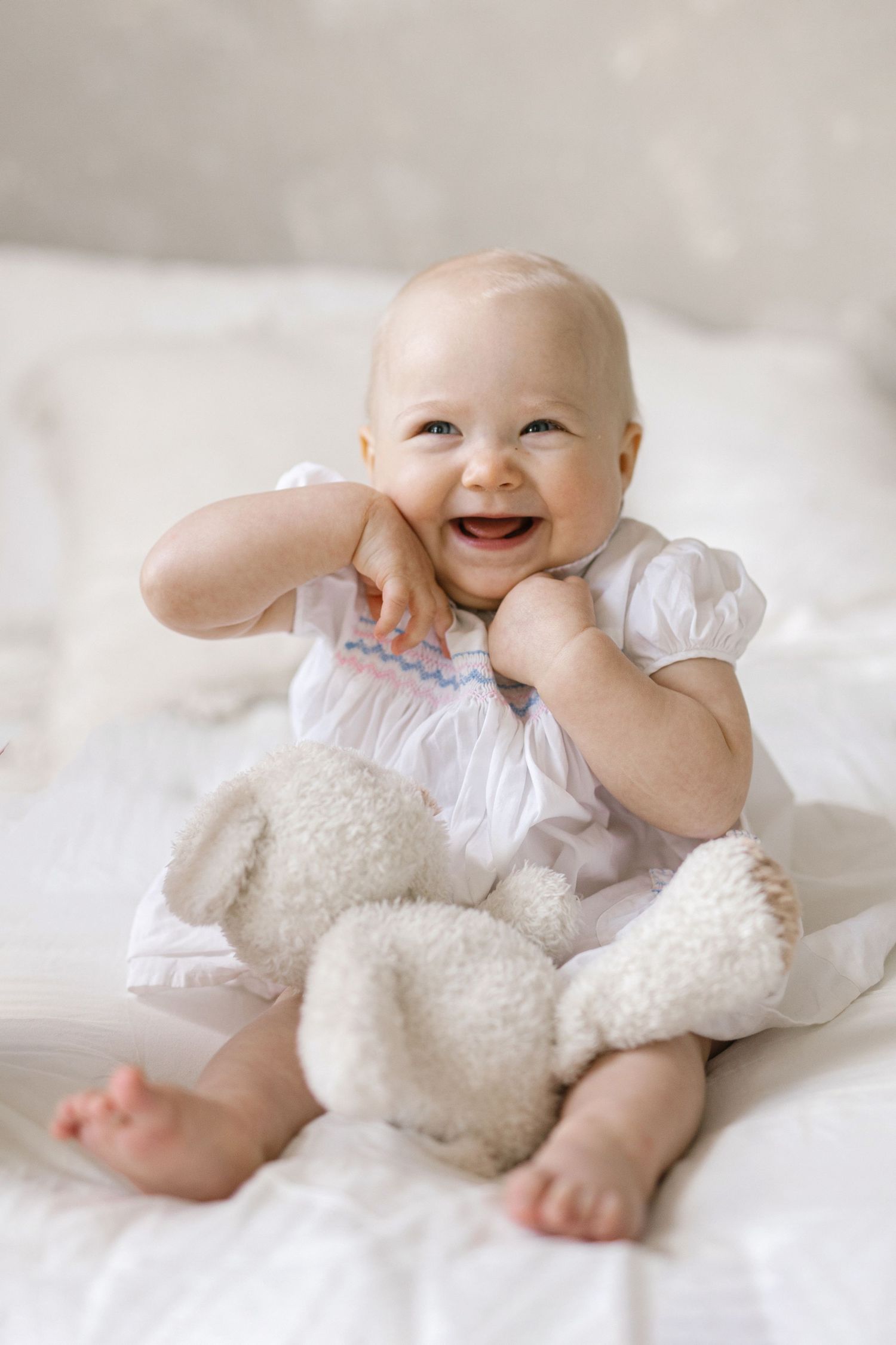 Smiling baby in white dress sitting on bed with stuffed animal toy, laughing joyfully in bright white nursery setting.