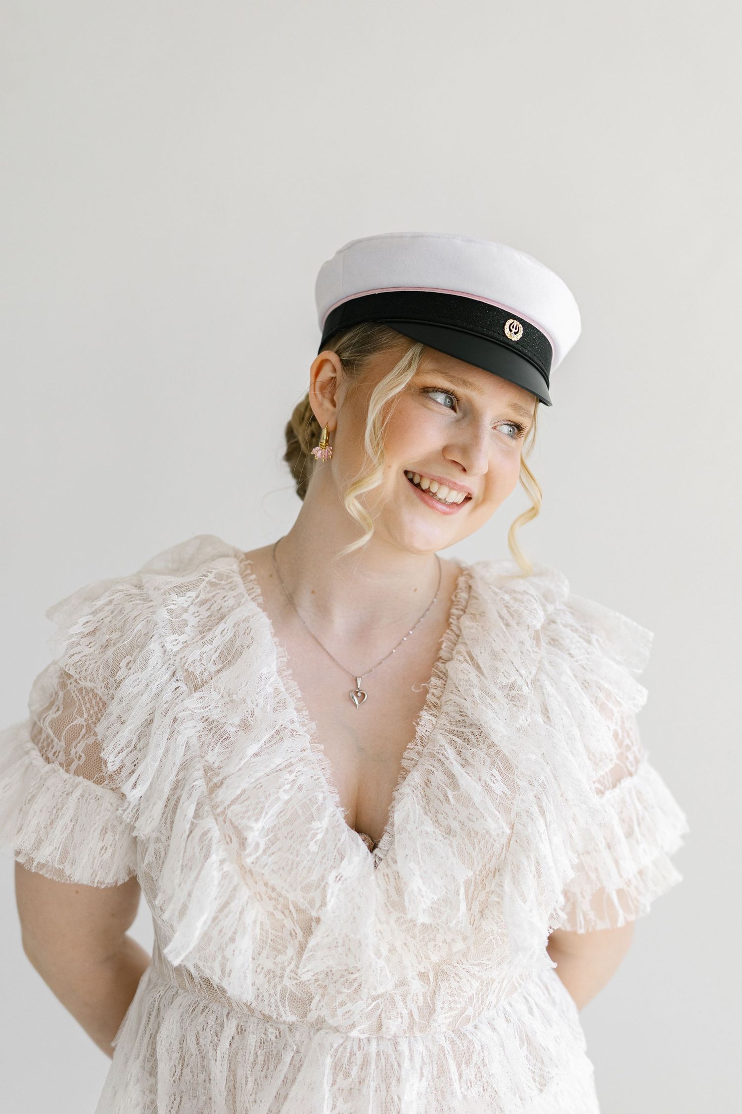 Smiling blonde woman in white lace dress and nautical cap poses against a light gray background.