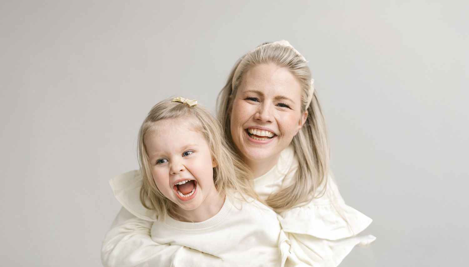 A joyful blonde woman and young girl with Down syndrome smile together, both wearing white outfits against a gray background.