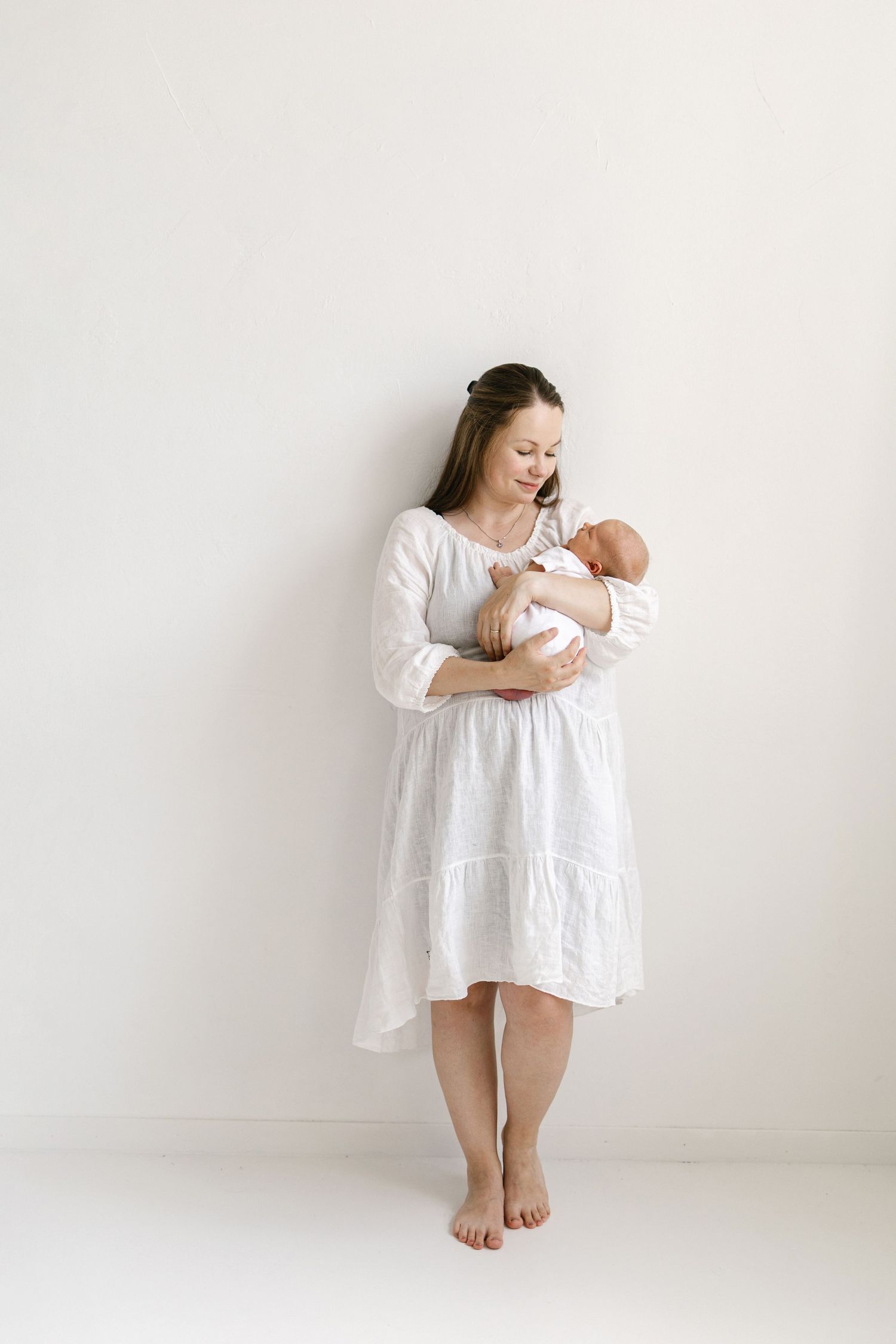 A barefoot mother in a white dress tenderly holds her newborn baby against a clean white background.