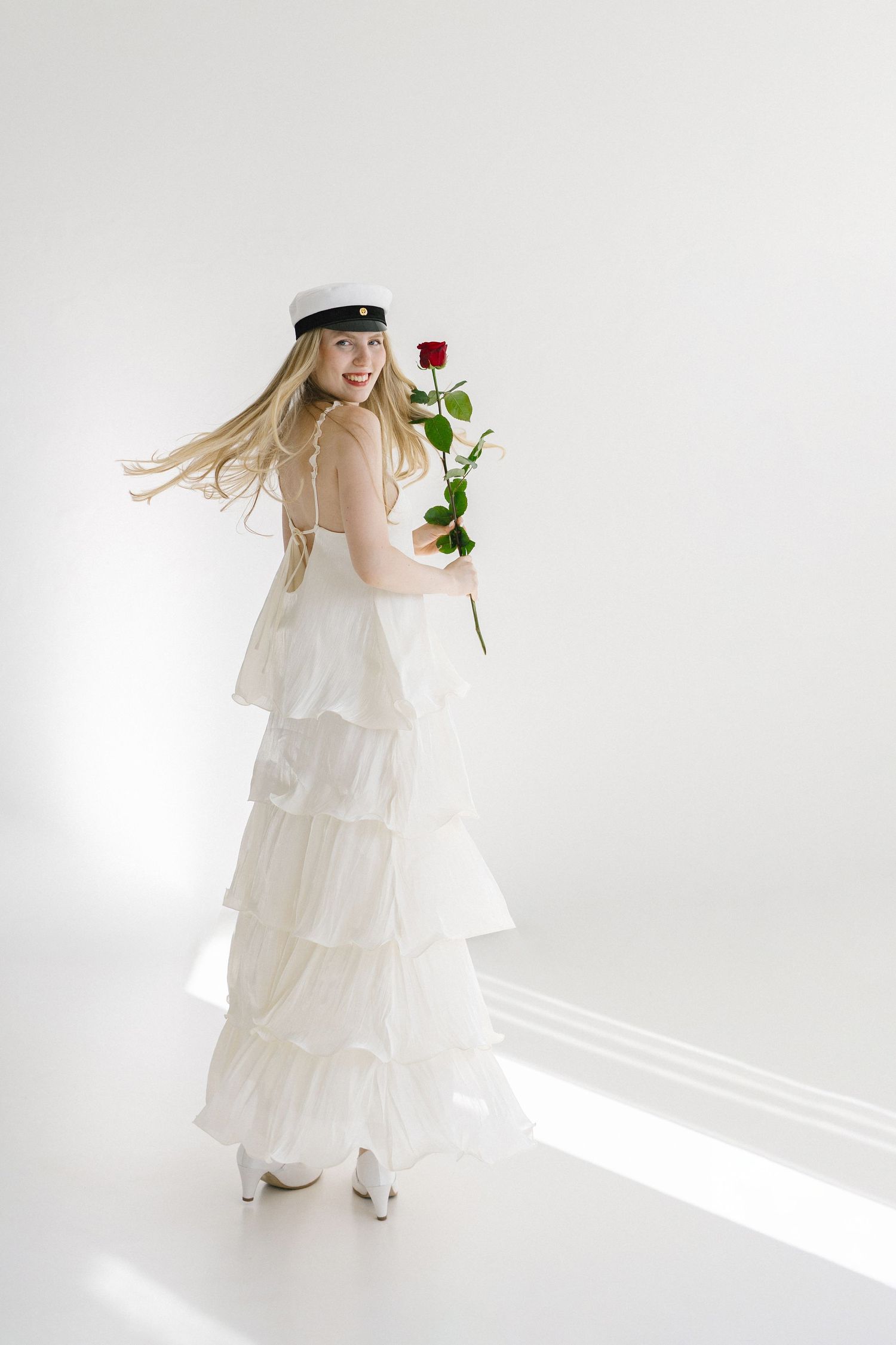 Young woman in white ruffled dress and sailor hat holding a red rose, posing elegantly in a bright studio.