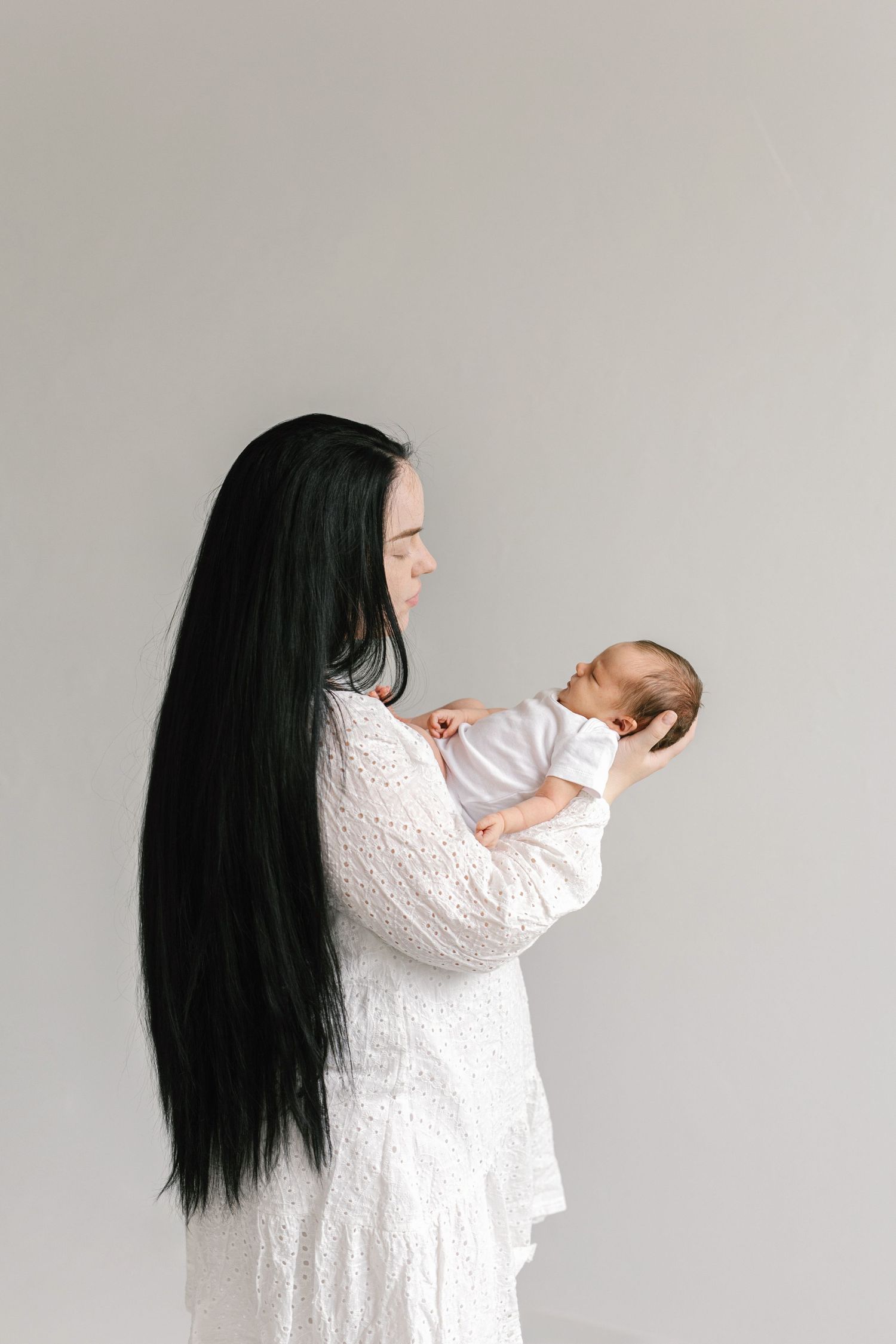 A mother with long dark hair in a white lace dress gently holds her newborn baby against a soft gray background.