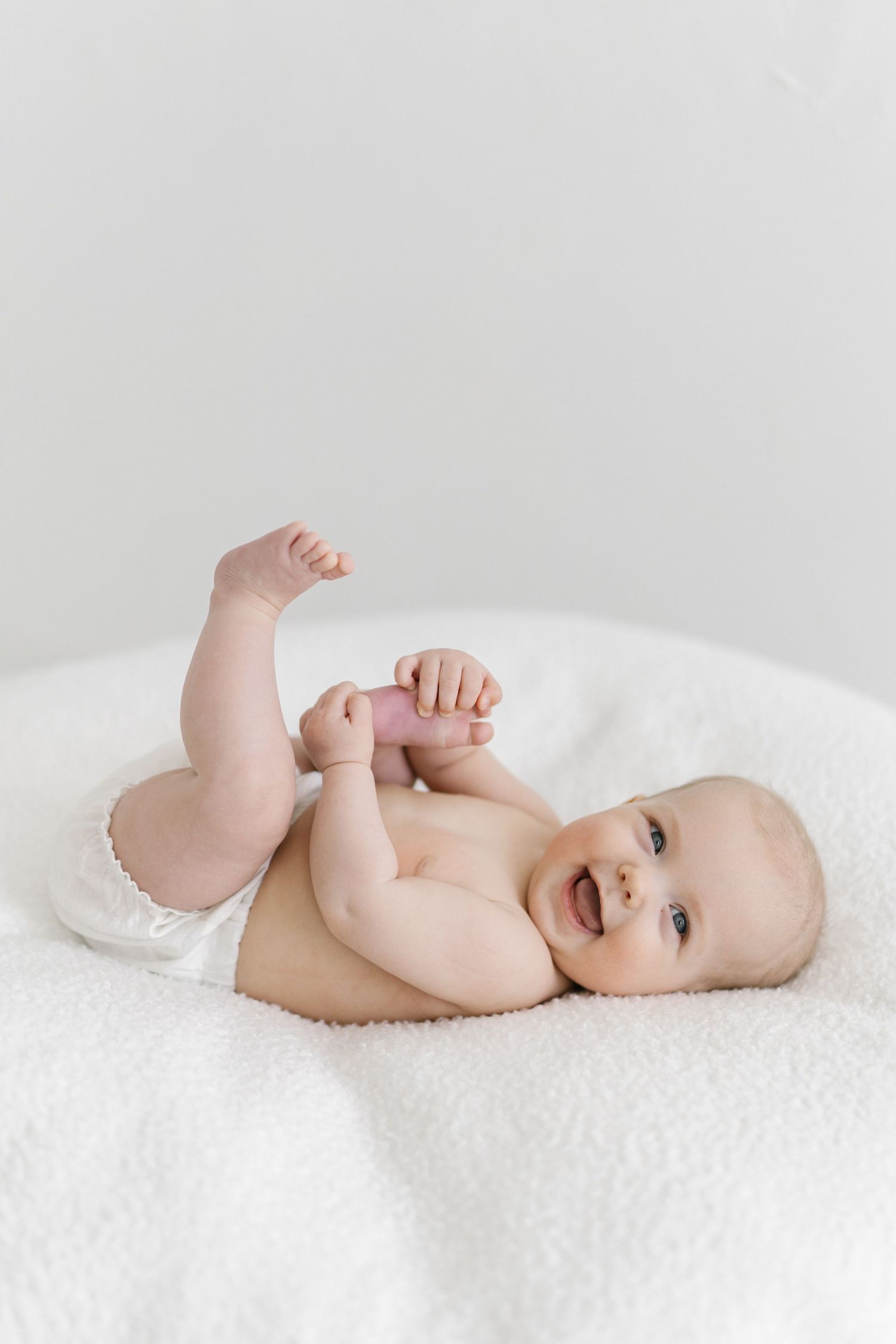 Smiling baby in a white diaper lying on a soft white blanket, reaching up with tiny hands and feet.