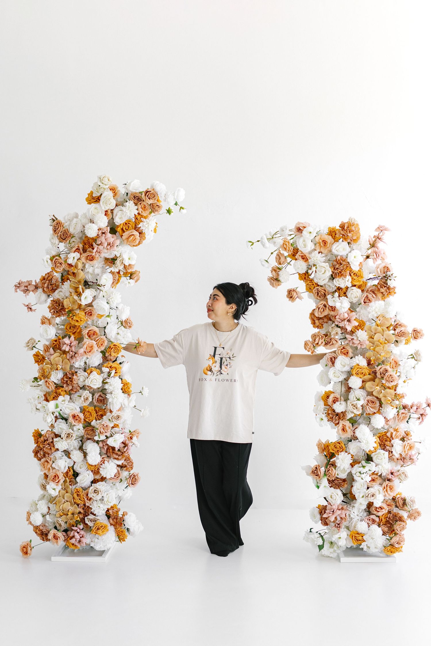 Person standing between two tall floral columns with orange, pink, and white roses on white background.