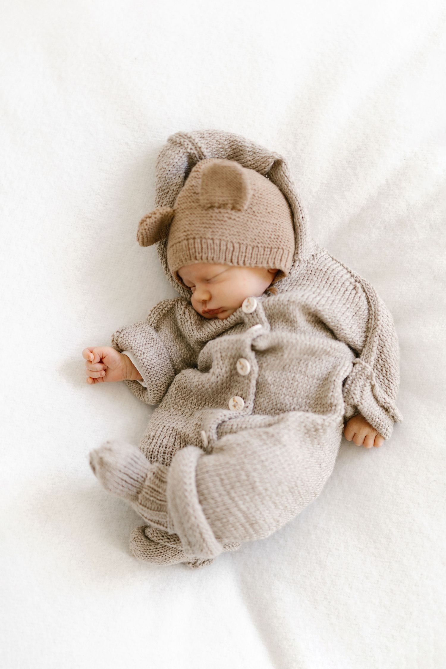 Sleeping newborn baby wearing a cozy beige knitted outfit with bear ear hat on a white blanket.