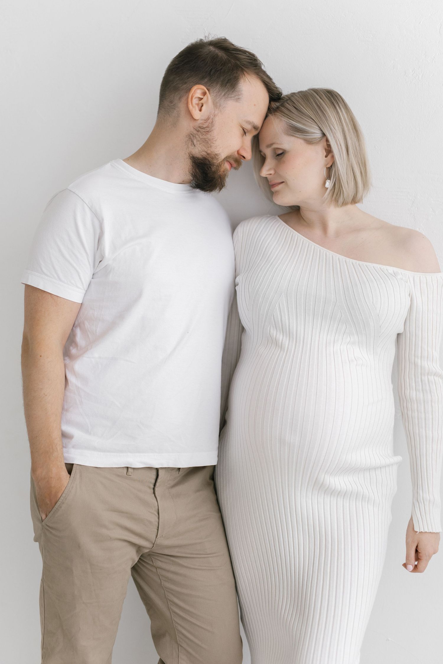 A pregnant woman in a white ribbed dress stands close with a bearded man in a white t-shirt, foreheads touching tenderly.