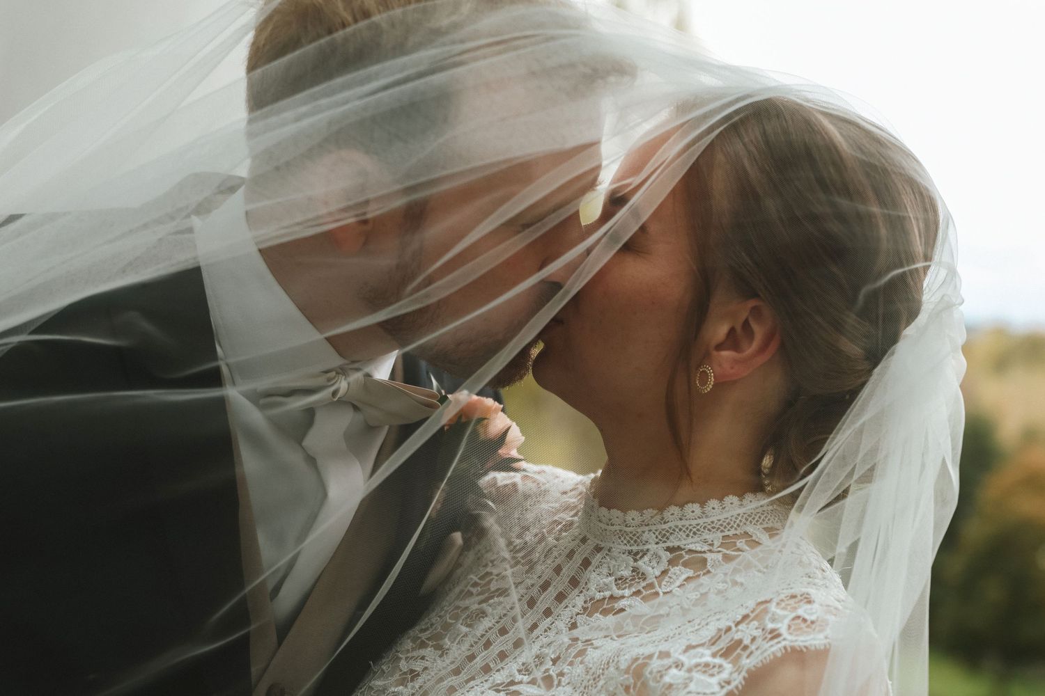 Bride and groom sharing a romantic kiss under a flowing white wedding veil outdoors.