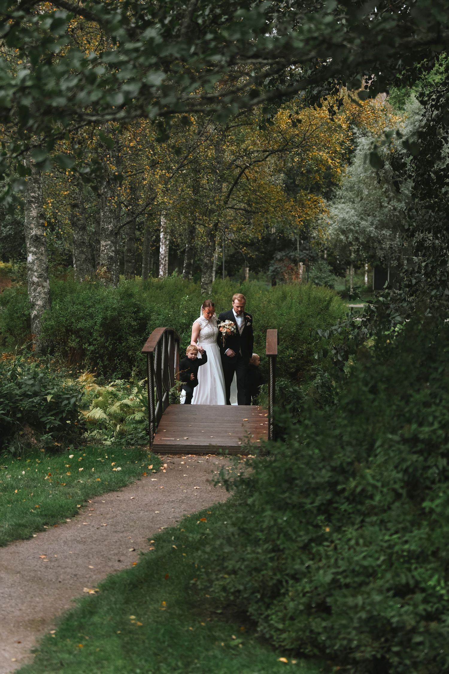 Bride and groom standing on a wooden bridge surrounded by lush green trees in a romantic garden wedding setting.
