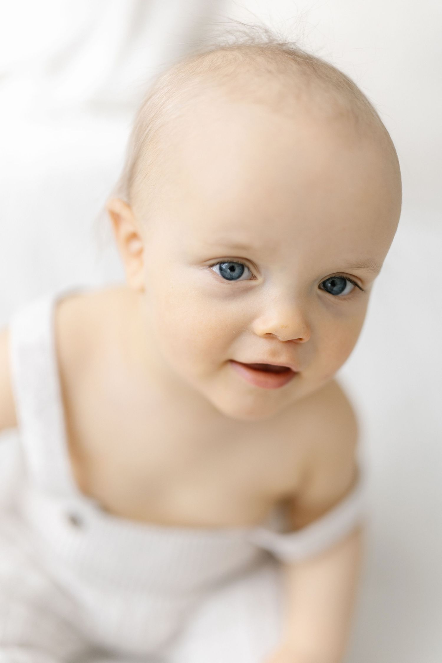 Adorable baby with blue eyes wearing a white romper, looking up with a sweet smile against a soft white background.