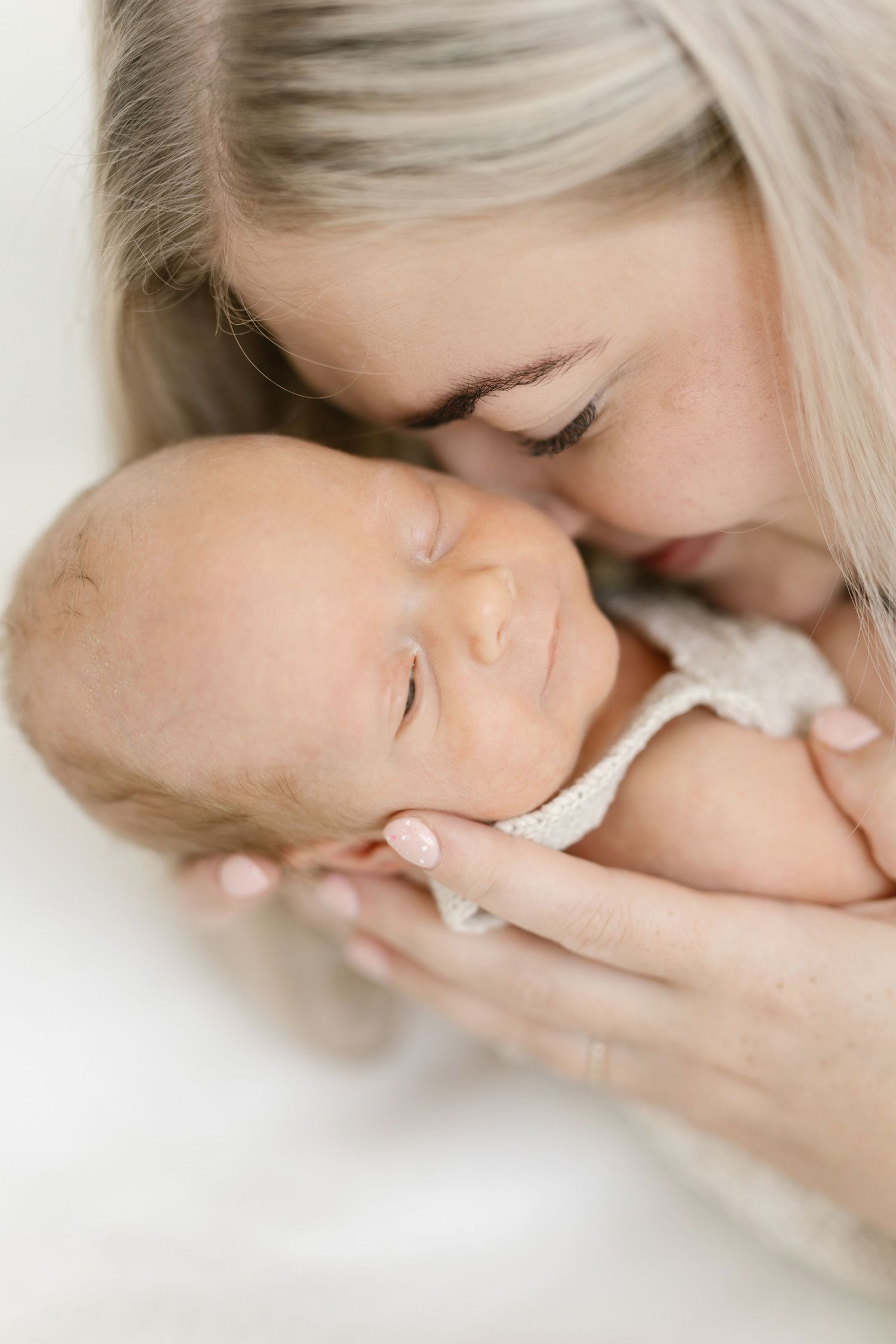 A mother with blonde hair gently kisses her sleeping newborn baby, cradling them tenderly in her hands.