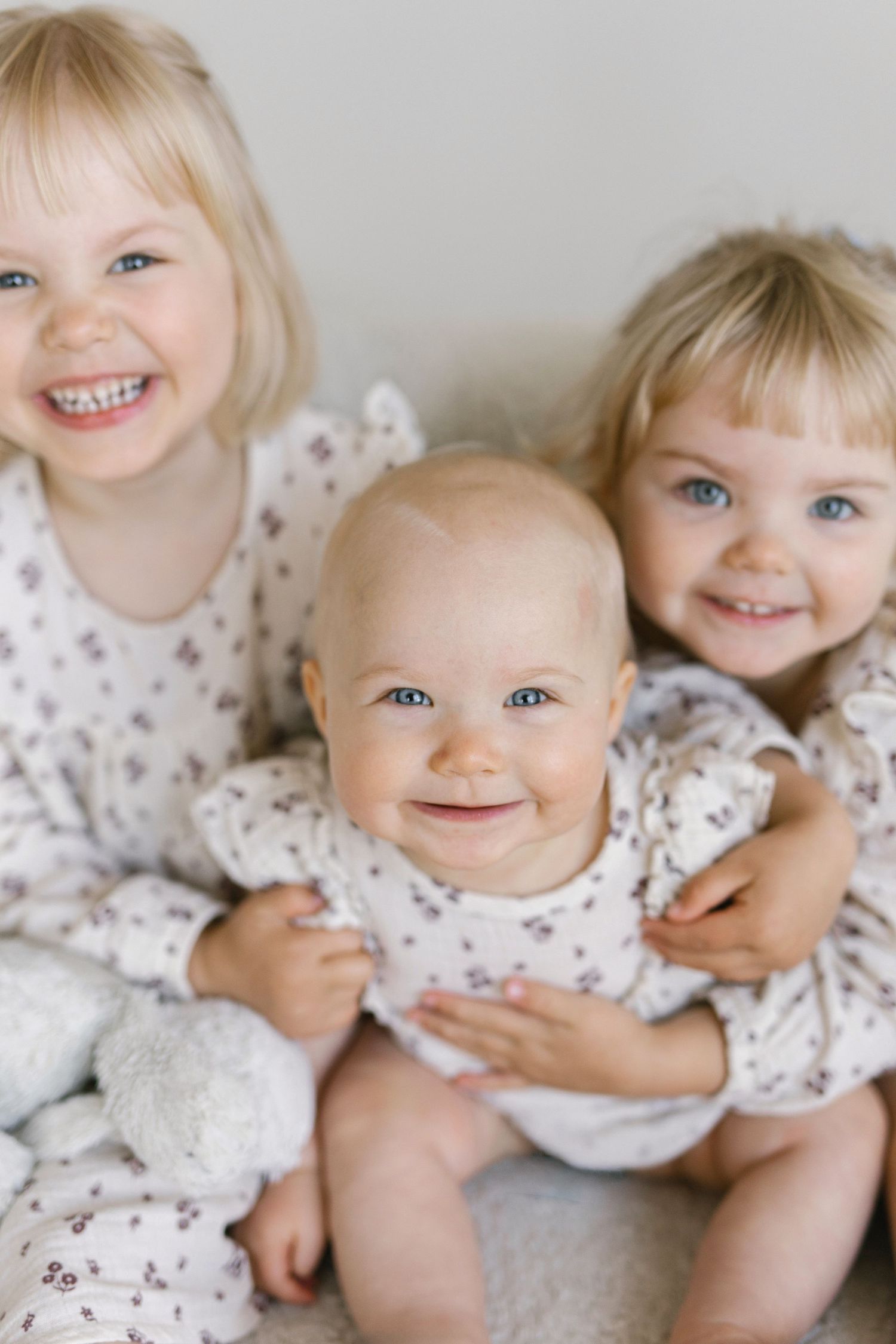 Three smiling blonde blue-eyed siblings in matching floral outfits, two toddlers hugging a happy baby.