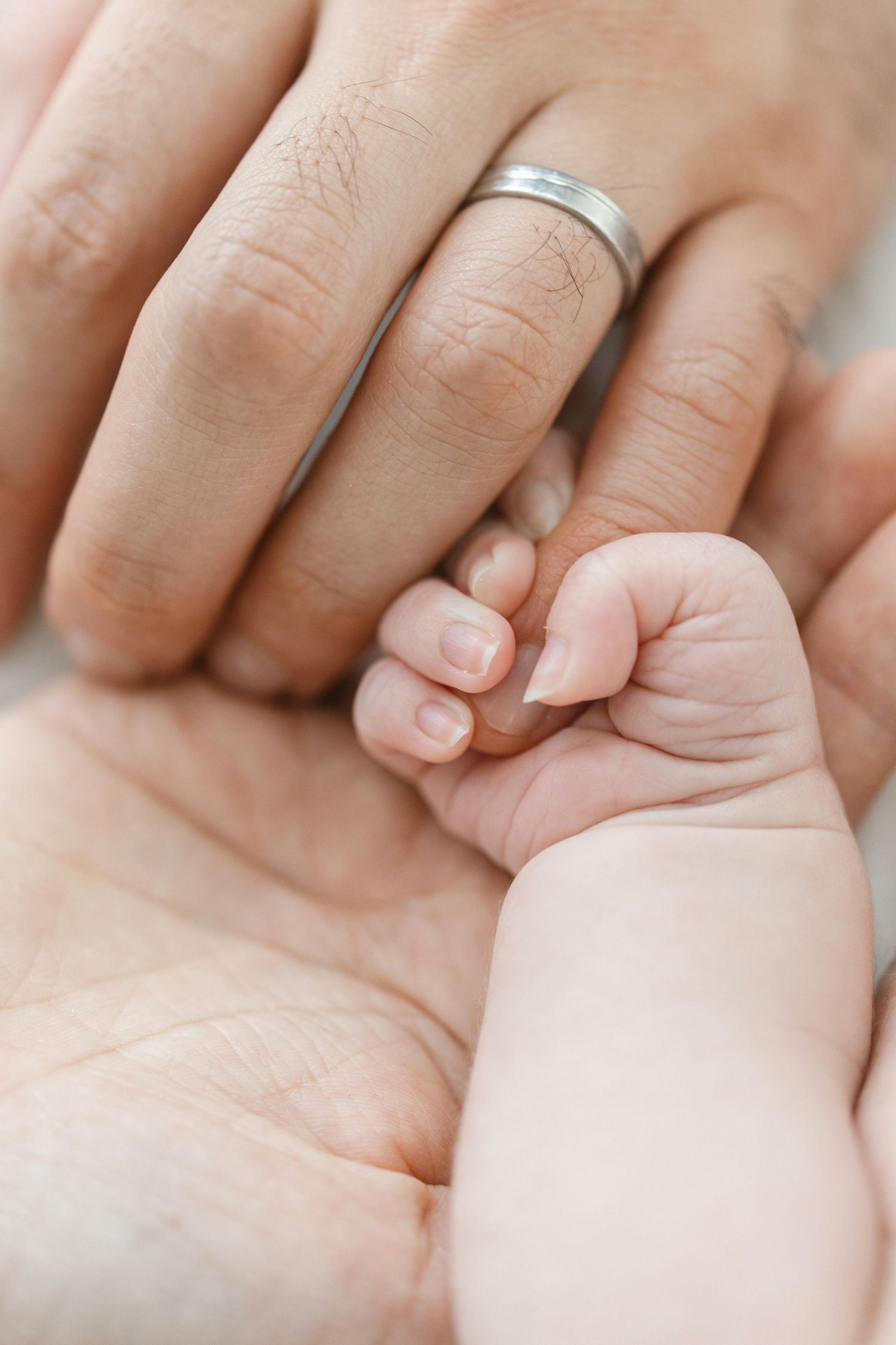 Newborn baby's tiny hand curled around an adult's finger, resting in a parent's open palm, with a silver wedding ring visible.
