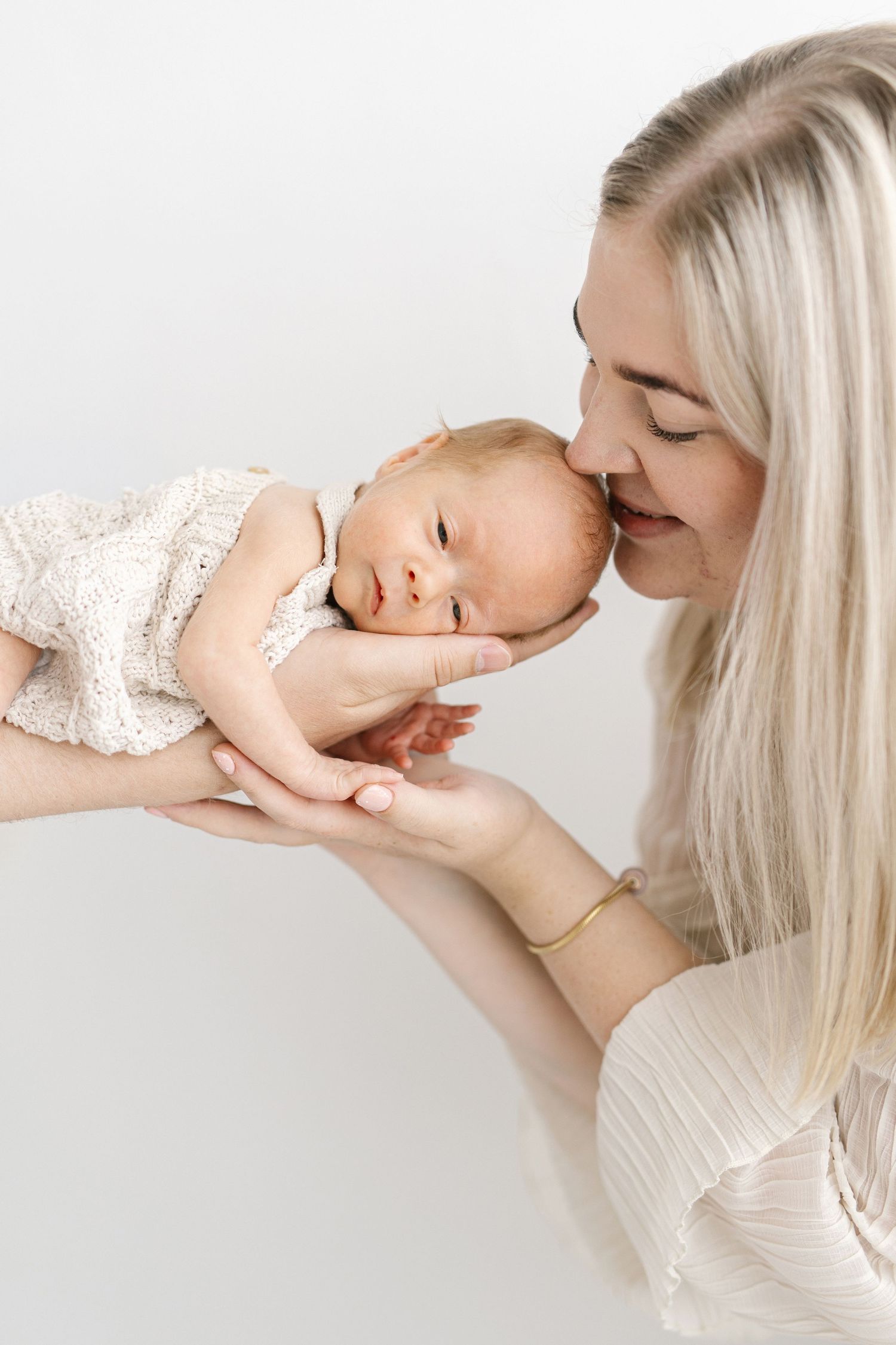 A blonde mother gently kisses her sleeping newborn baby dressed in white, held tenderly in her hands.