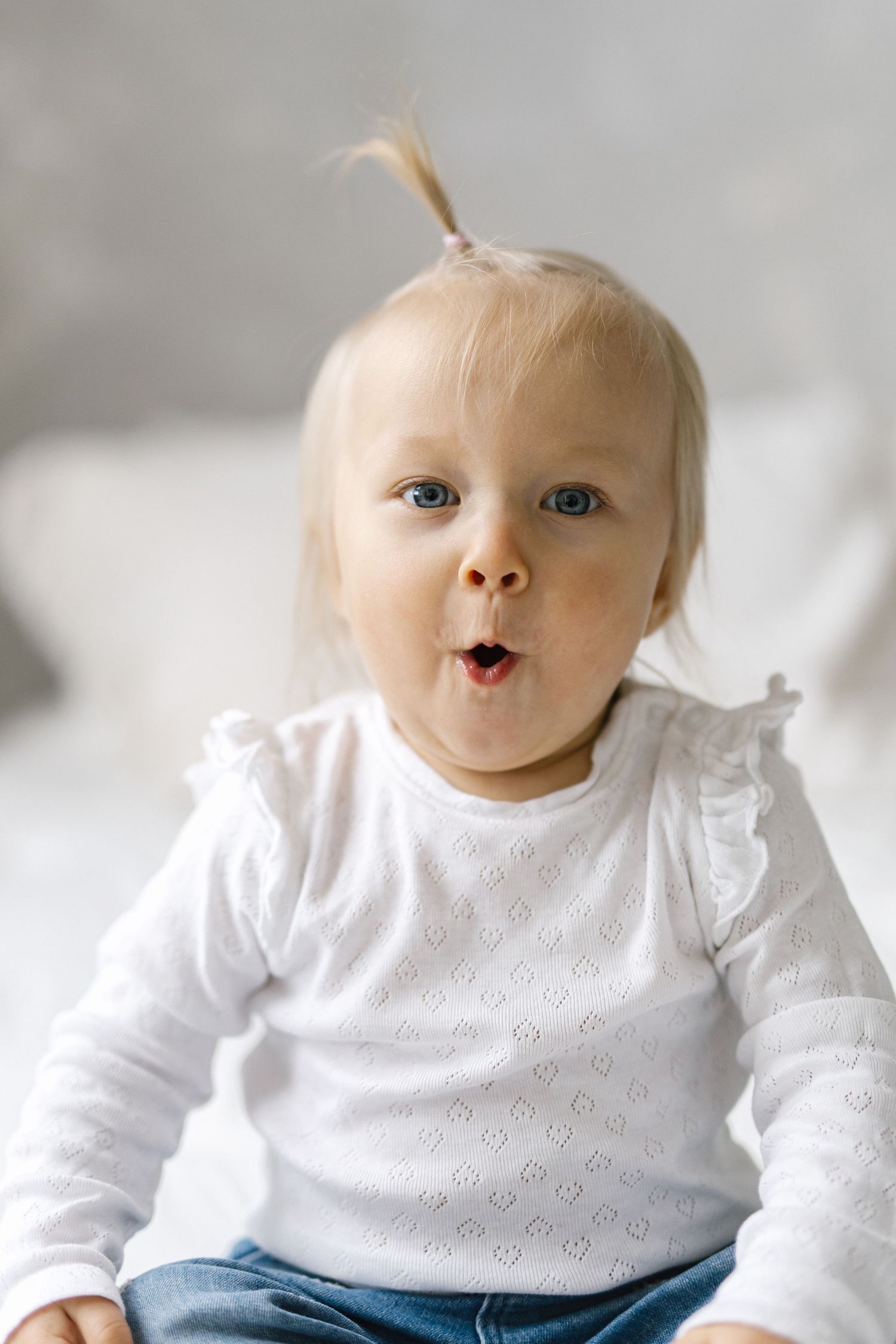 Adorable blonde toddler girl in white top and jeans making a funny surprised face while sitting indoors.