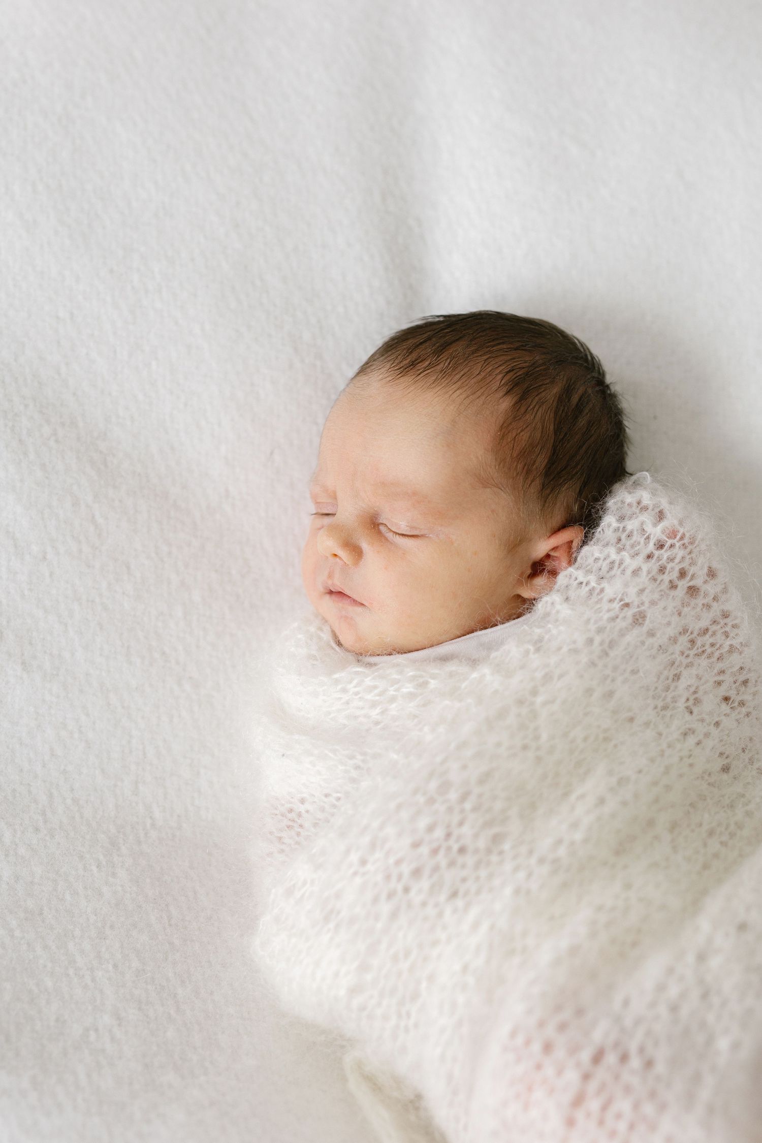 Peaceful newborn baby sleeping swaddled in a soft white knit wrap on a white blanket.