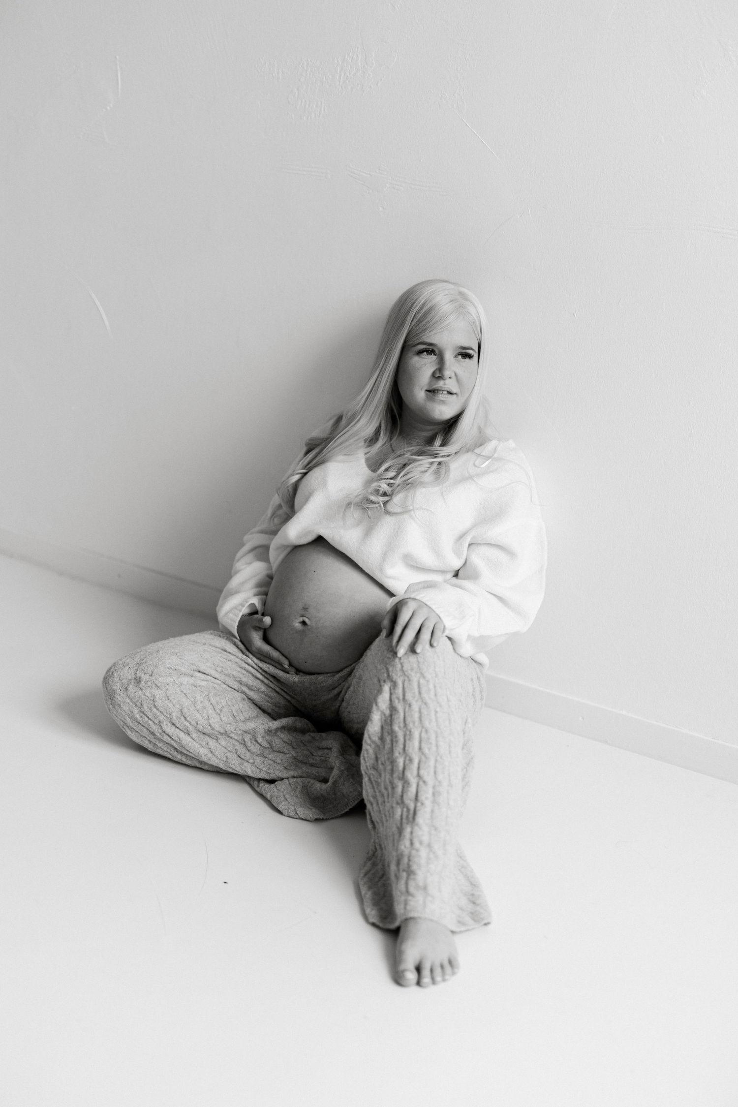 Black and white maternity photo of a blonde woman sitting on floor cradling her baby bump in cozy knit pants and crop sweater.