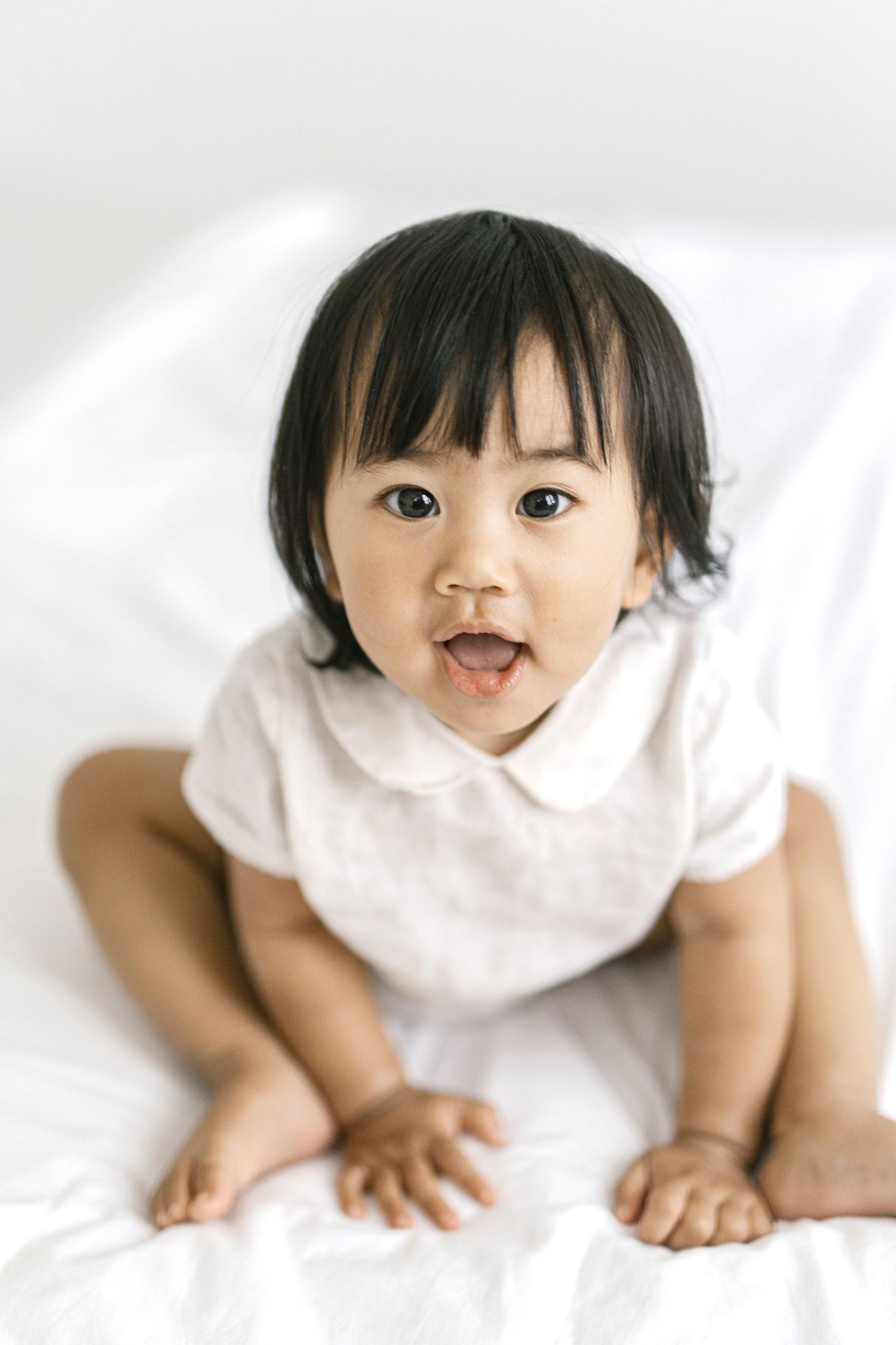 Adorable Asian baby in white romper sitting on white bed, looking up with bright eyes and open mouth smile.