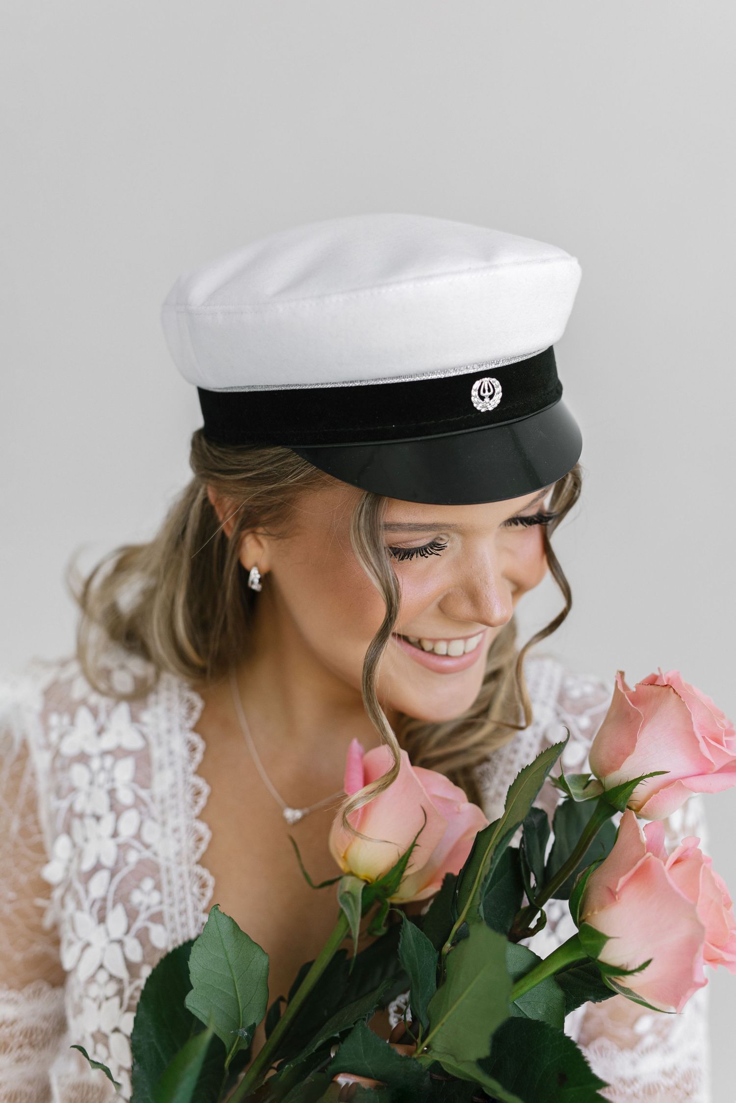 Smiling bride in white lace dress and sailor cap holding pink roses bouquet on light gray background.