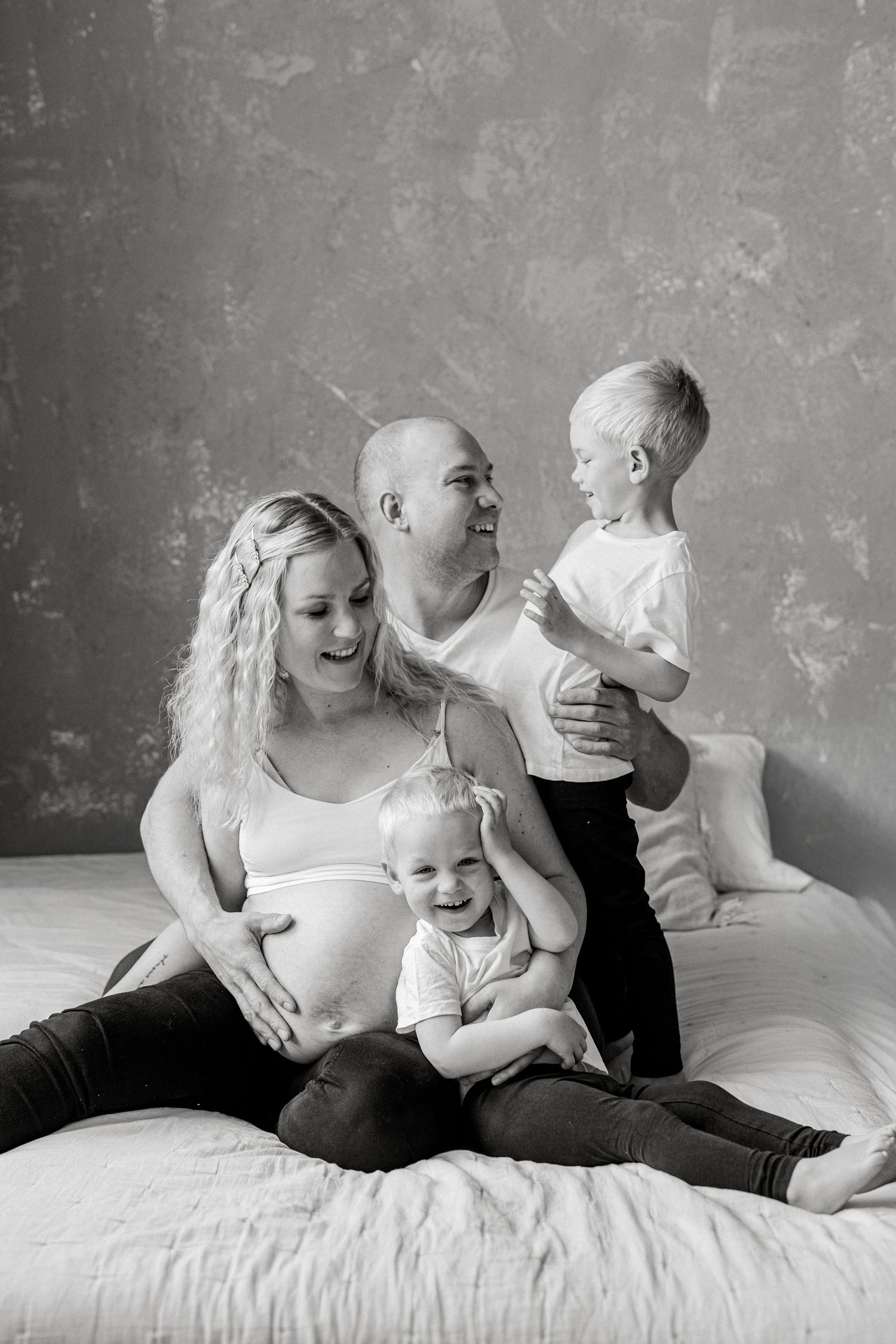 Black and white family portrait of pregnant woman, bald man, and two young children sitting together on a bed.