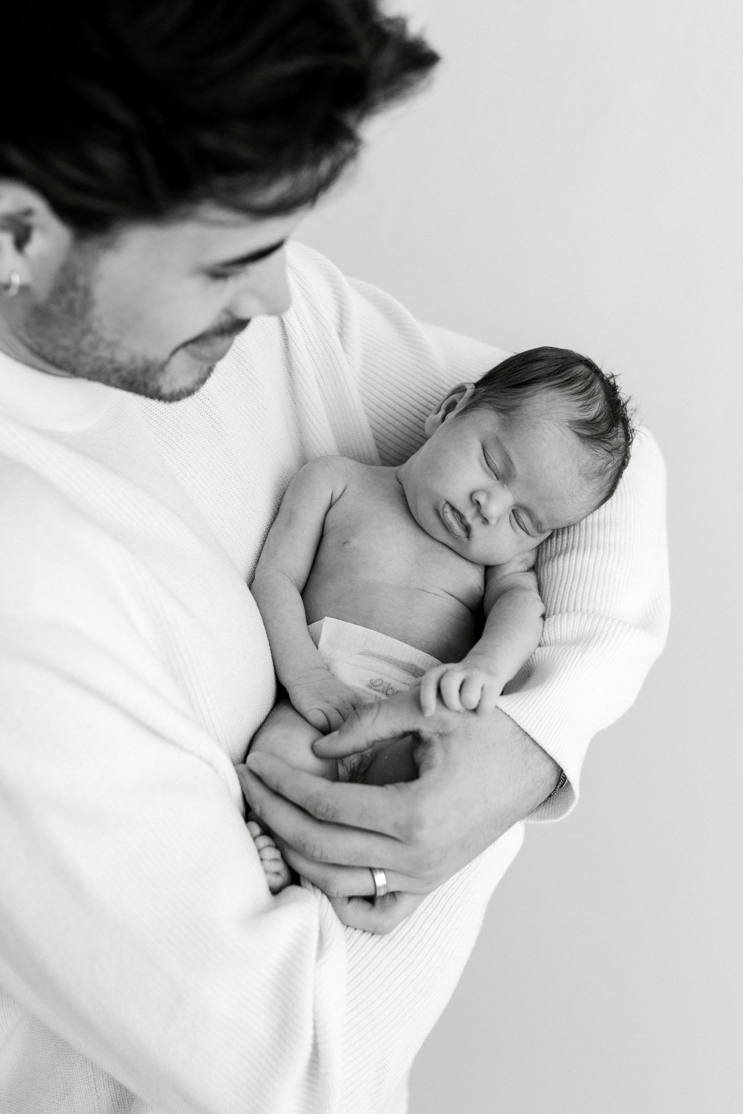 Black and white photo of a father cradling his sleeping newborn baby wrapped in a soft white blanket.