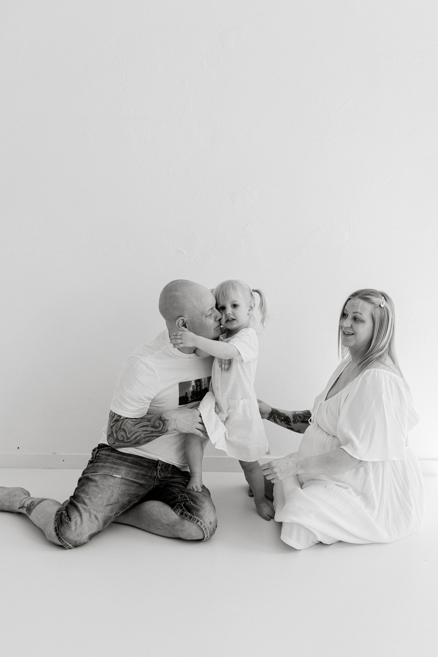 Black and white photo of a tattooed bald man and blonde woman sitting on floor with their toddler daughter in white dress.