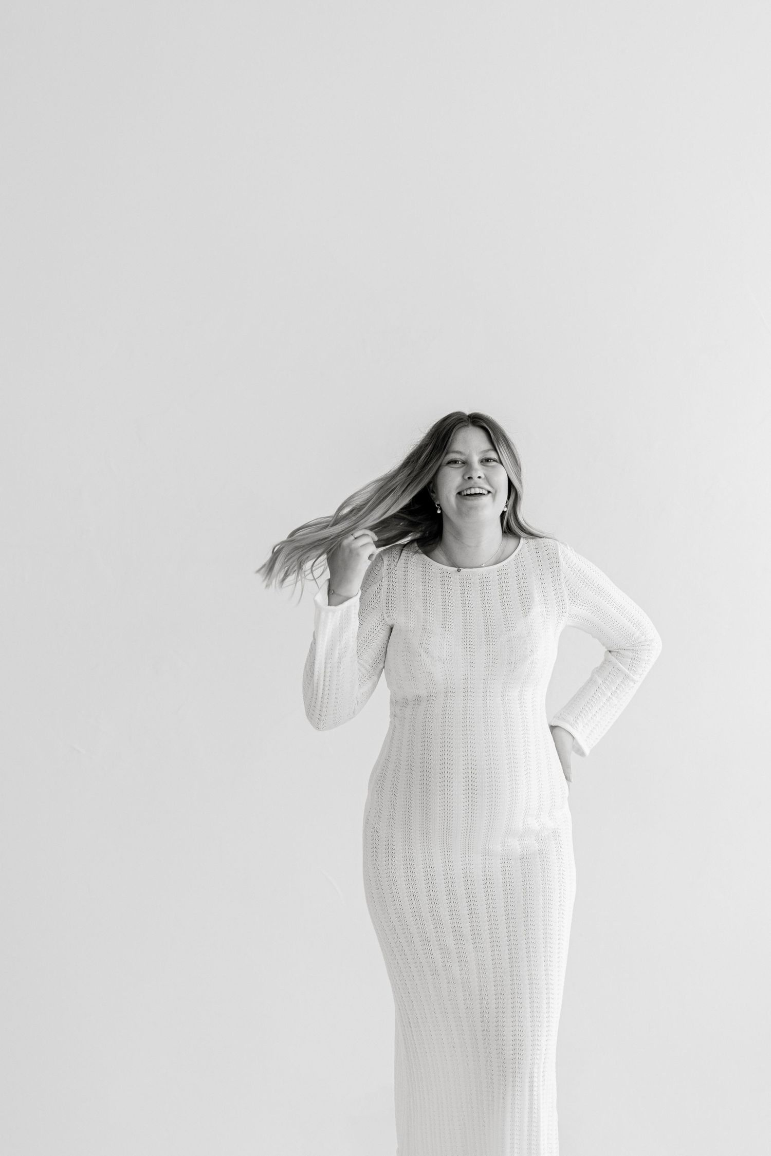 A smiling woman in a white ribbed dress playfully tosses her hair against a light background.