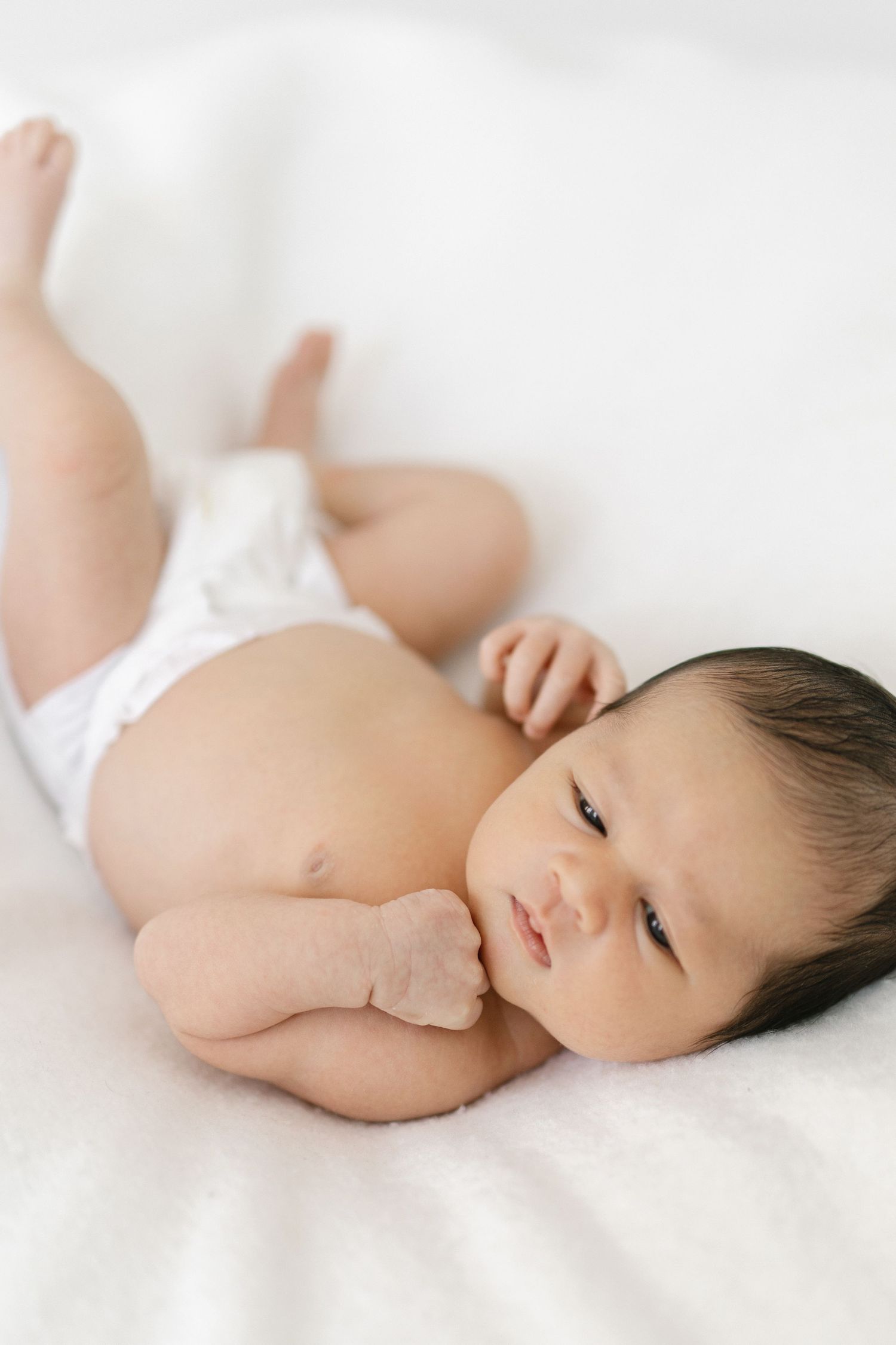 A newborn baby with dark hair lies on a white blanket wearing a diaper, gazing softly with tiny hands near face.