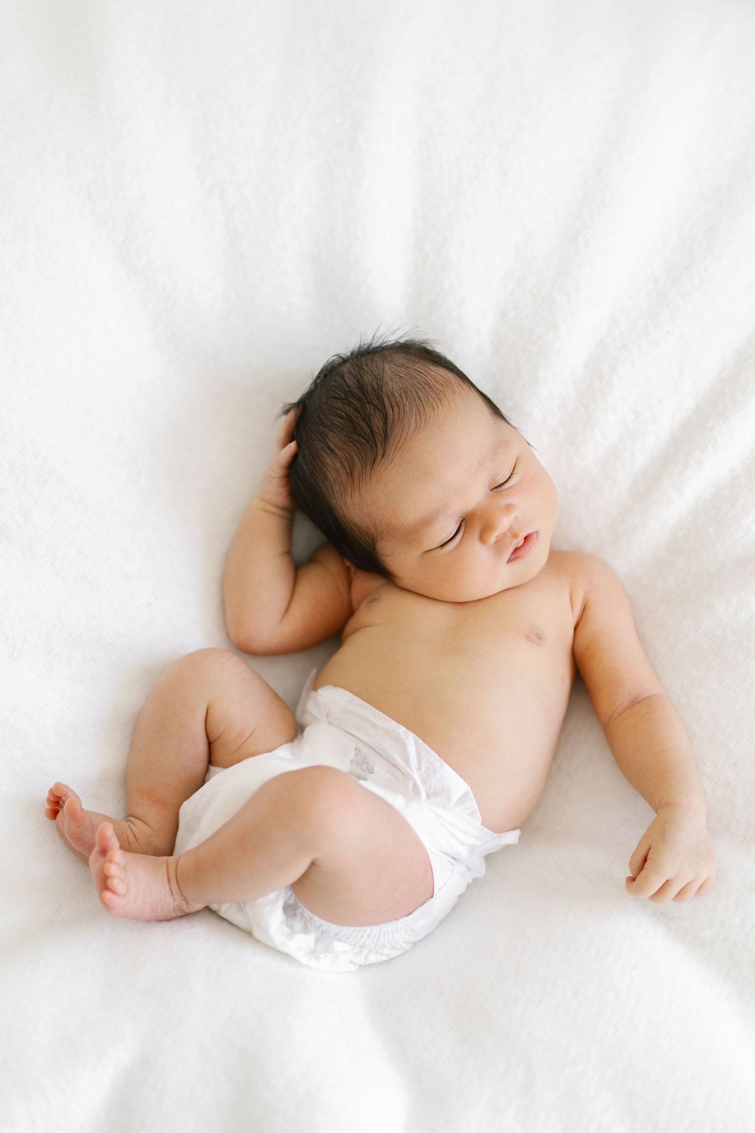 Peaceful newborn baby sleeping on white blanket wearing diaper with dark hair and relaxed arms.