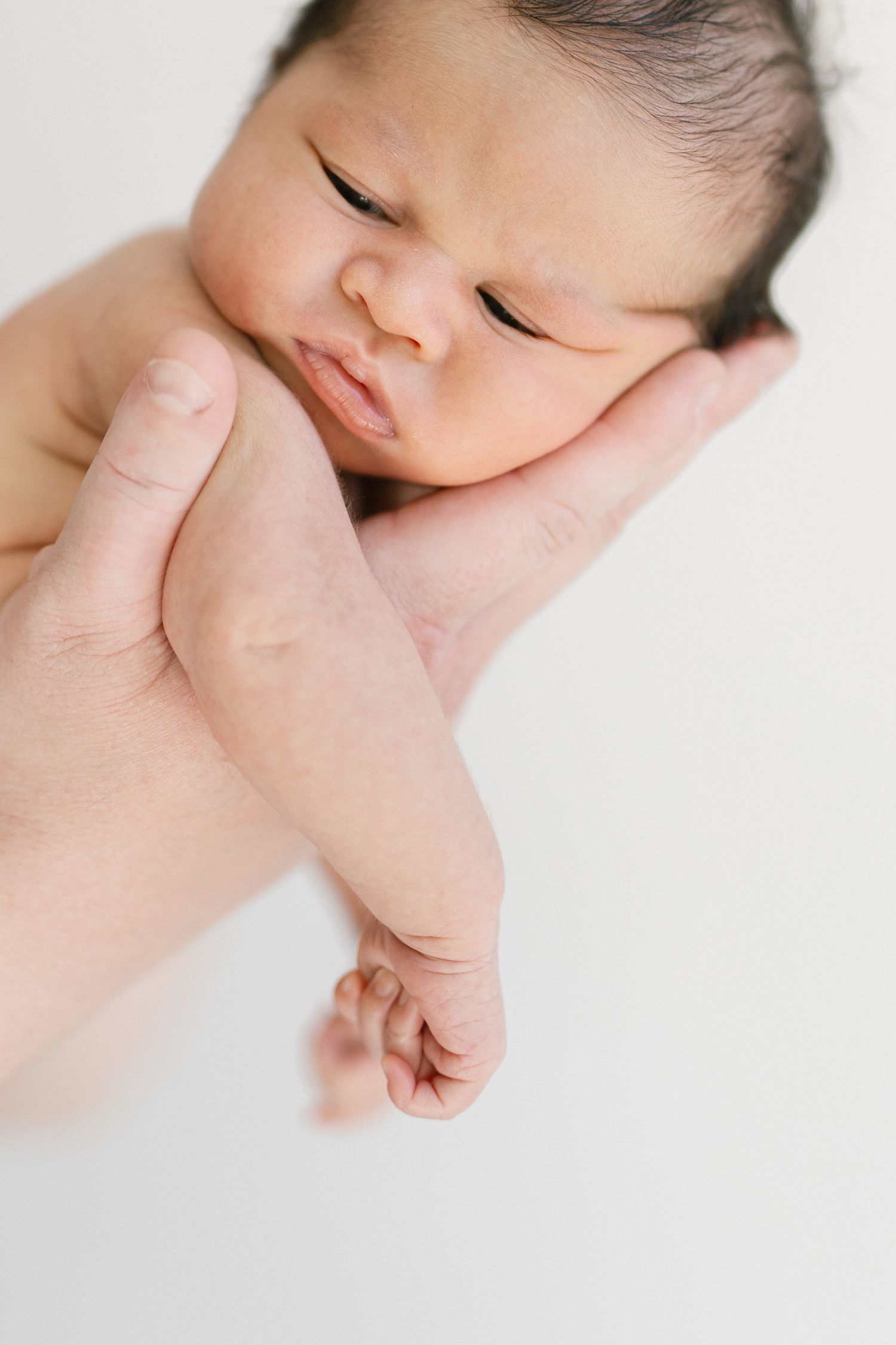 Newborn baby resting peacefully in parent's hand, with dark hair and tiny fist, against white background.