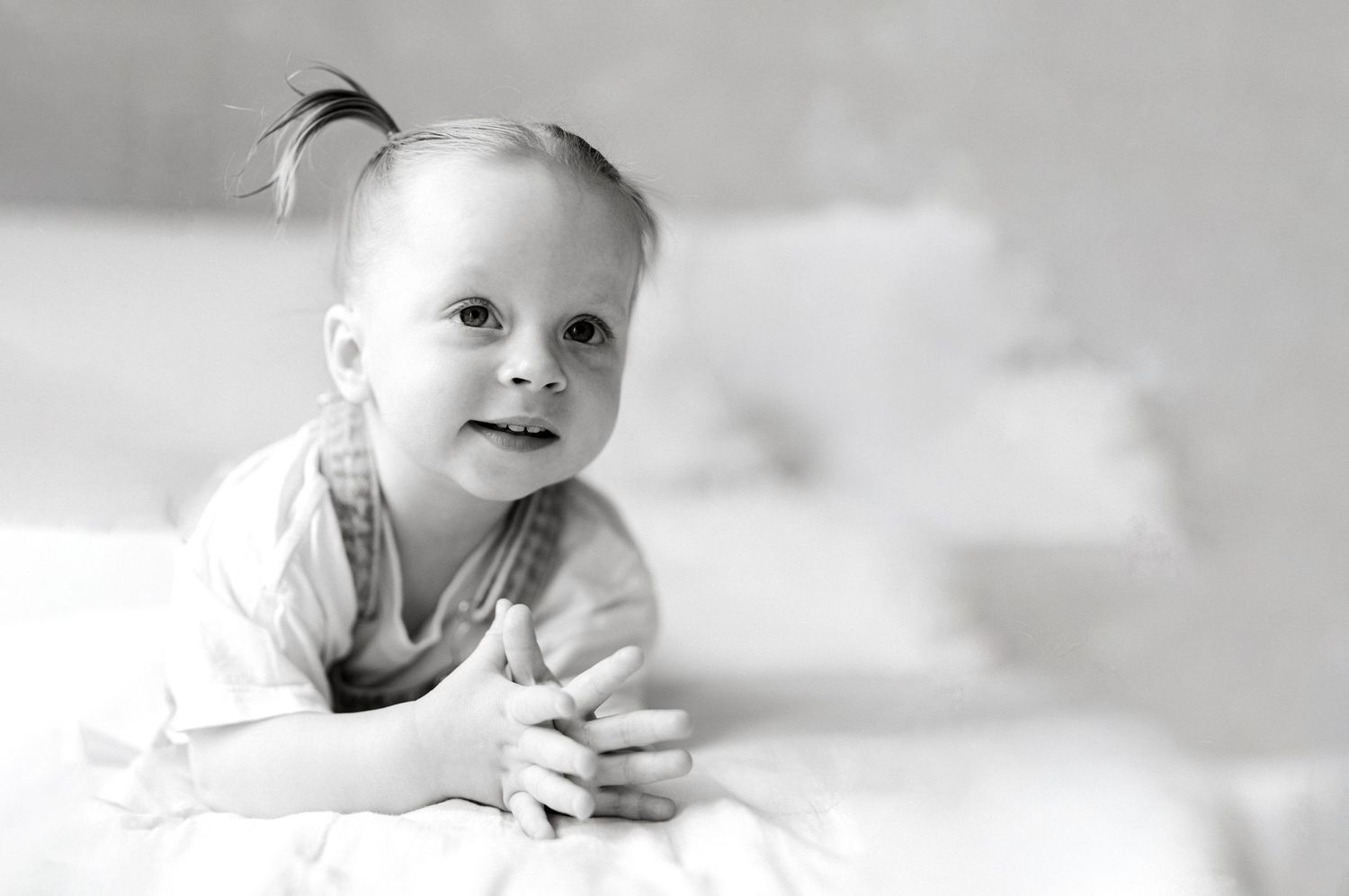 Black and white photo of a smiling baby girl with a tiny ponytail lying on her tummy on a white bed.