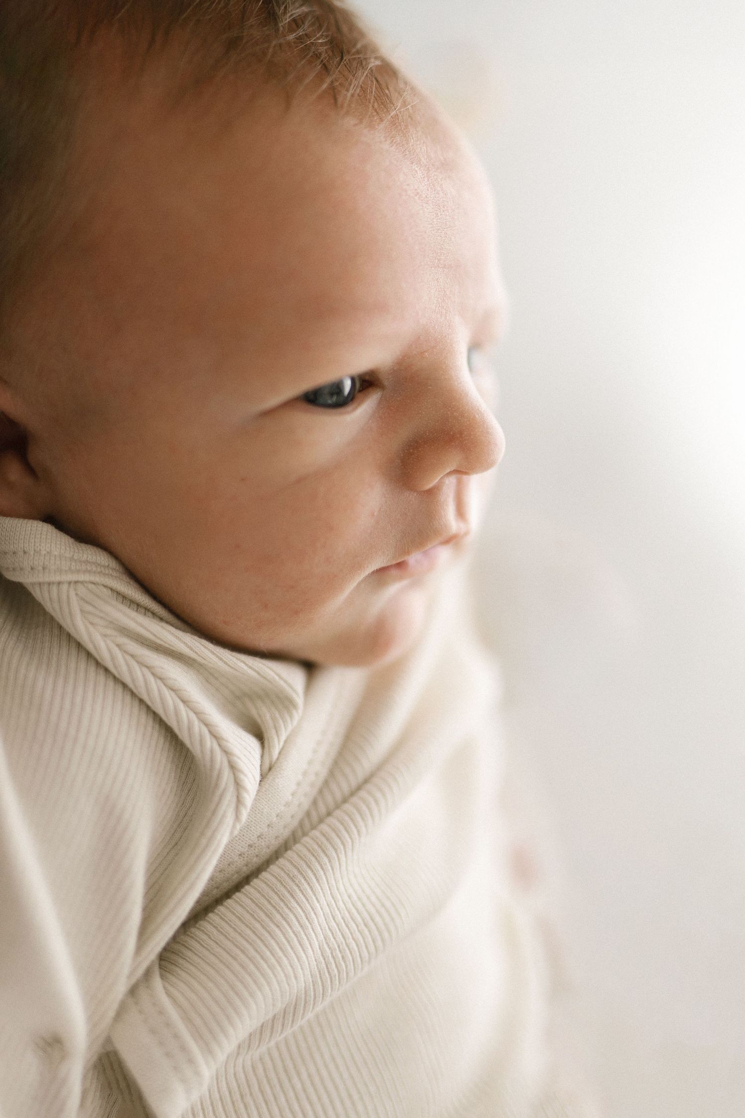 Close-up portrait of a newborn baby in a cream ribbed wrap, gazing sideways with bright blue eyes.