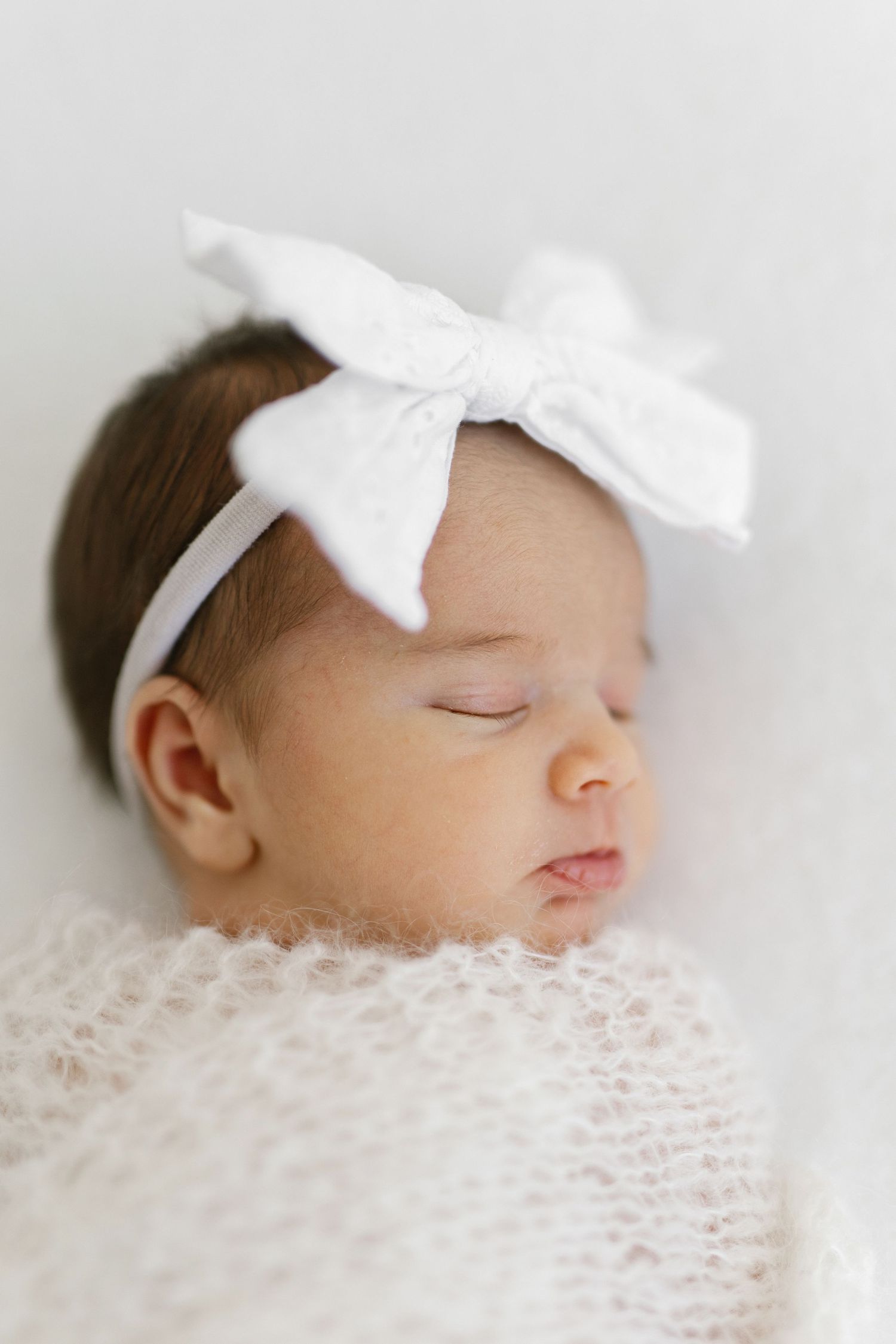 Sleeping newborn baby wearing white bow headband wrapped in soft white lace blanket.