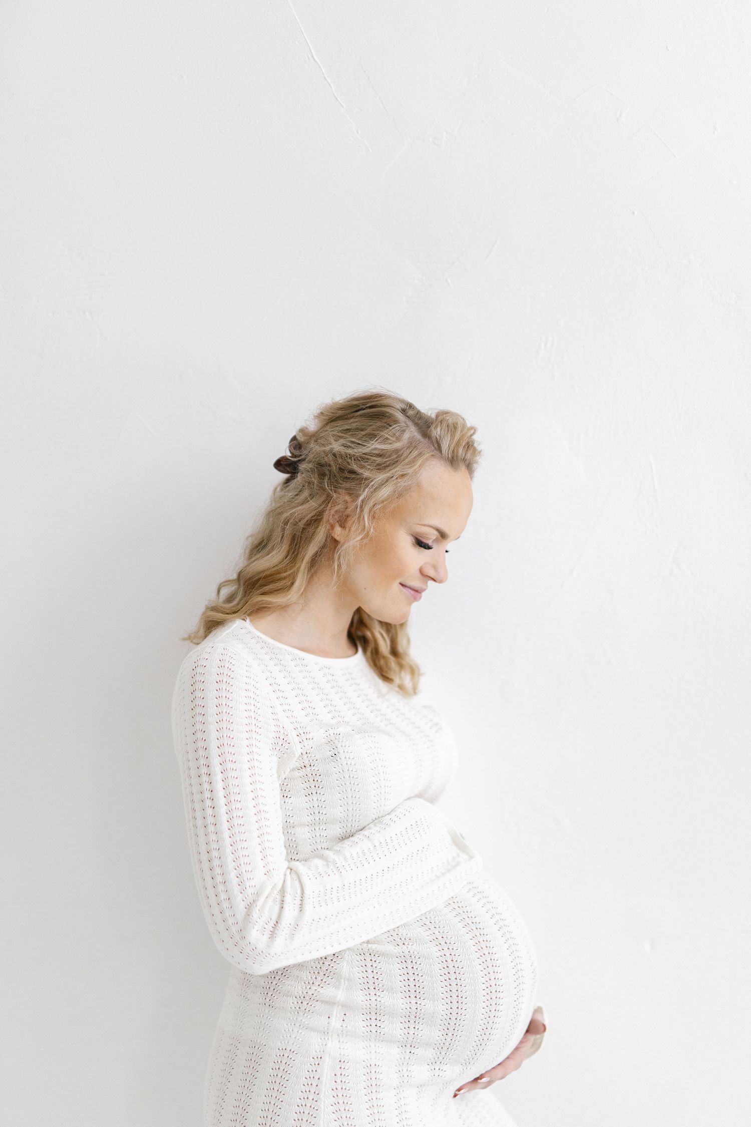 Elegant maternity portrait in white knit dress against minimalist white background.