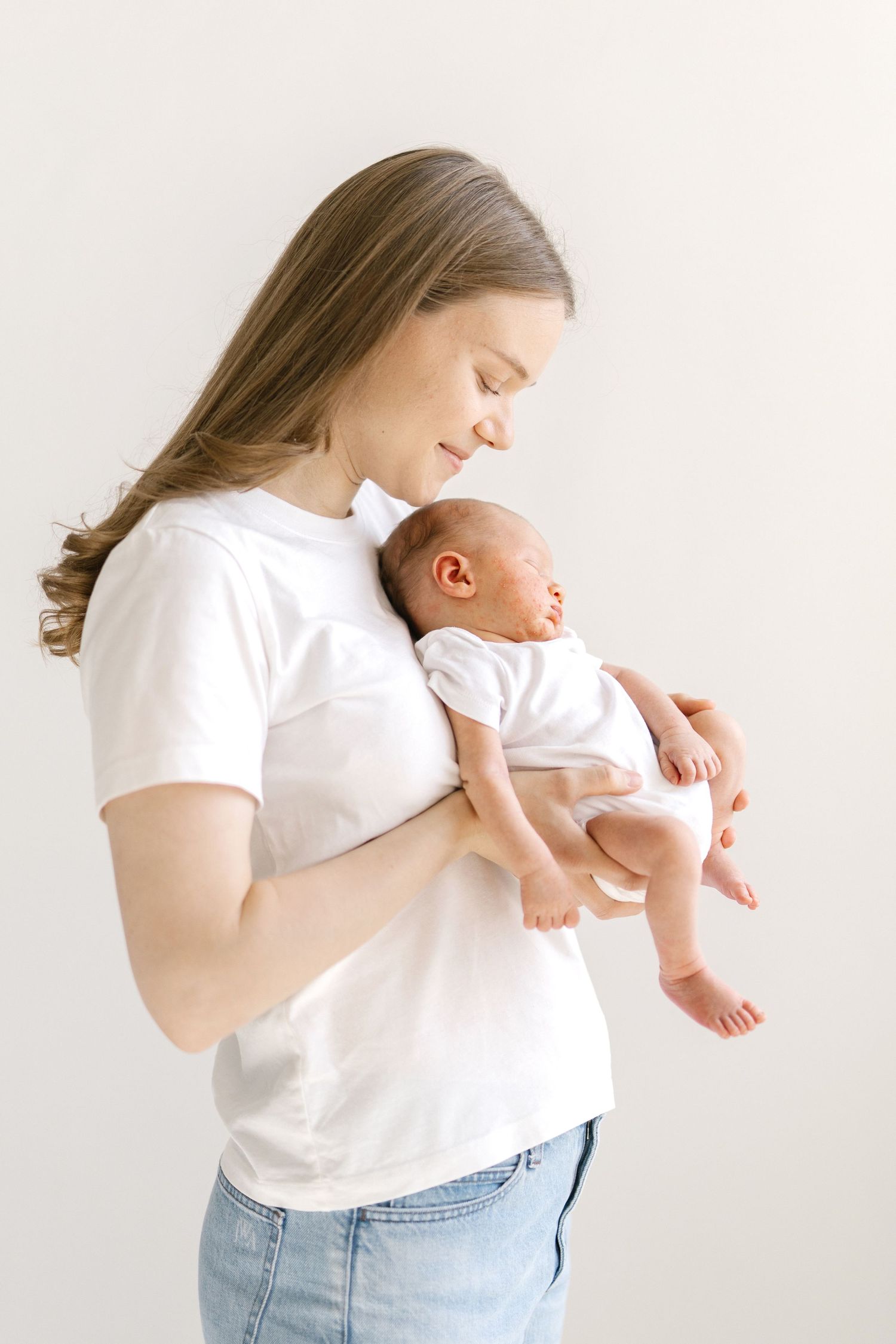 Young mother in white t-shirt tenderly holds sleeping newborn baby against light background.