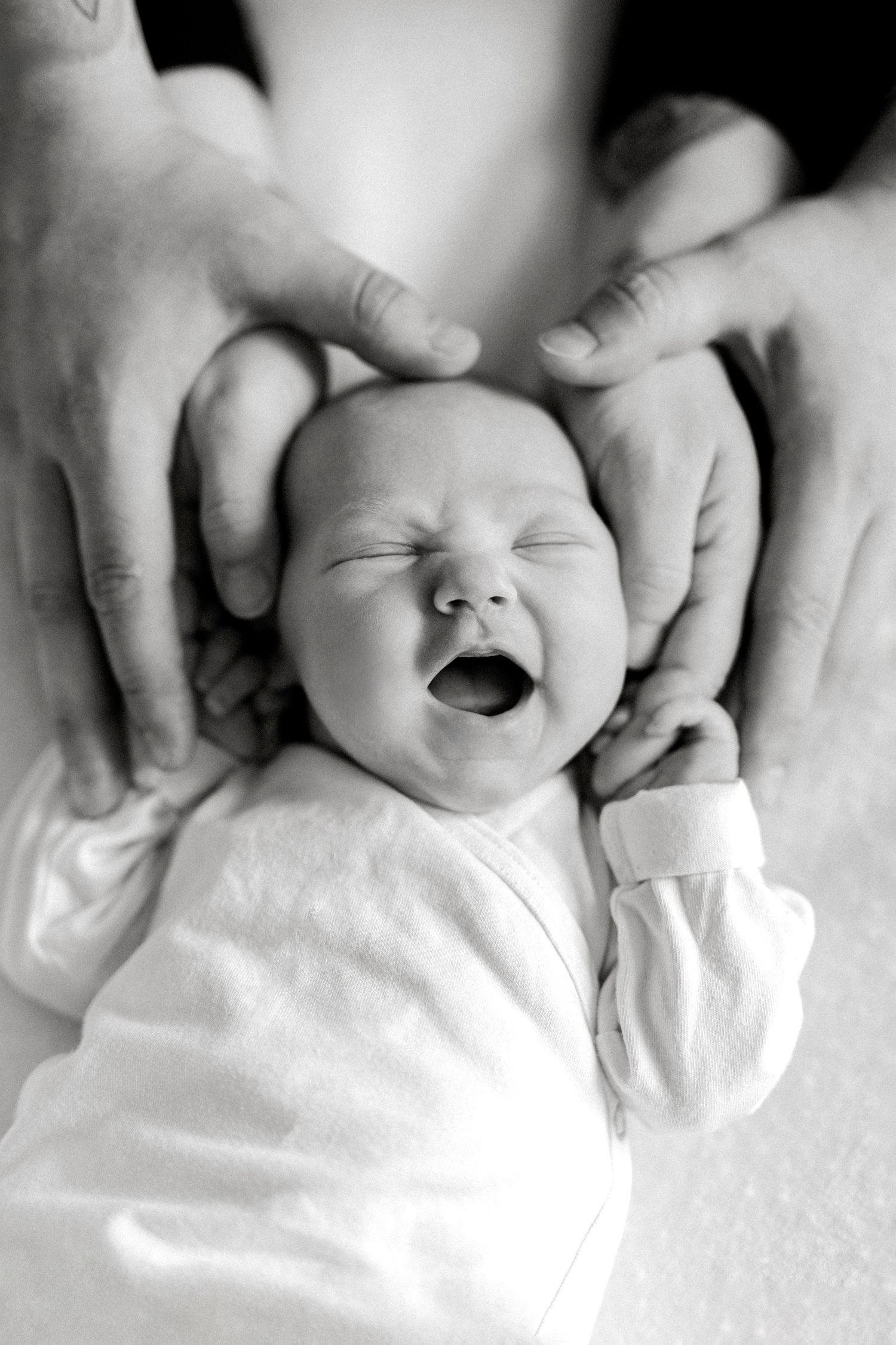 Black and white photo of a yawning newborn baby cradled gently in loving parents hands.