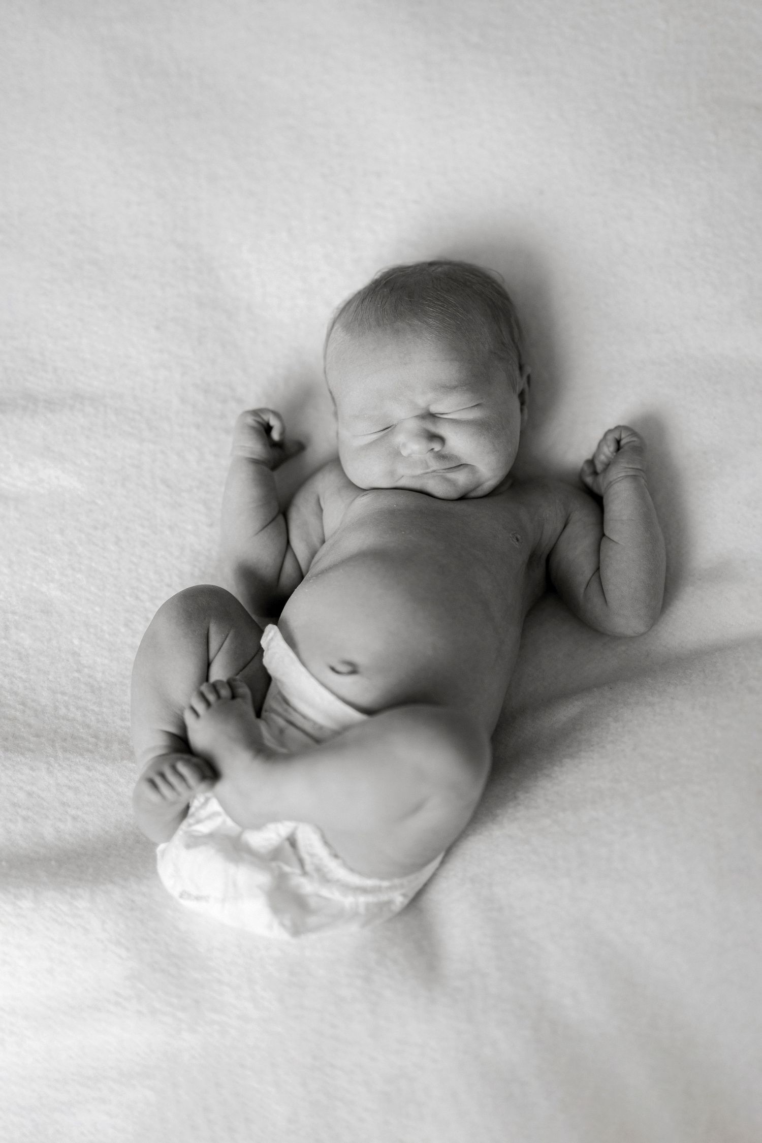 Black and white photo of a sleeping newborn baby lying on a soft white blanket in a diaper.