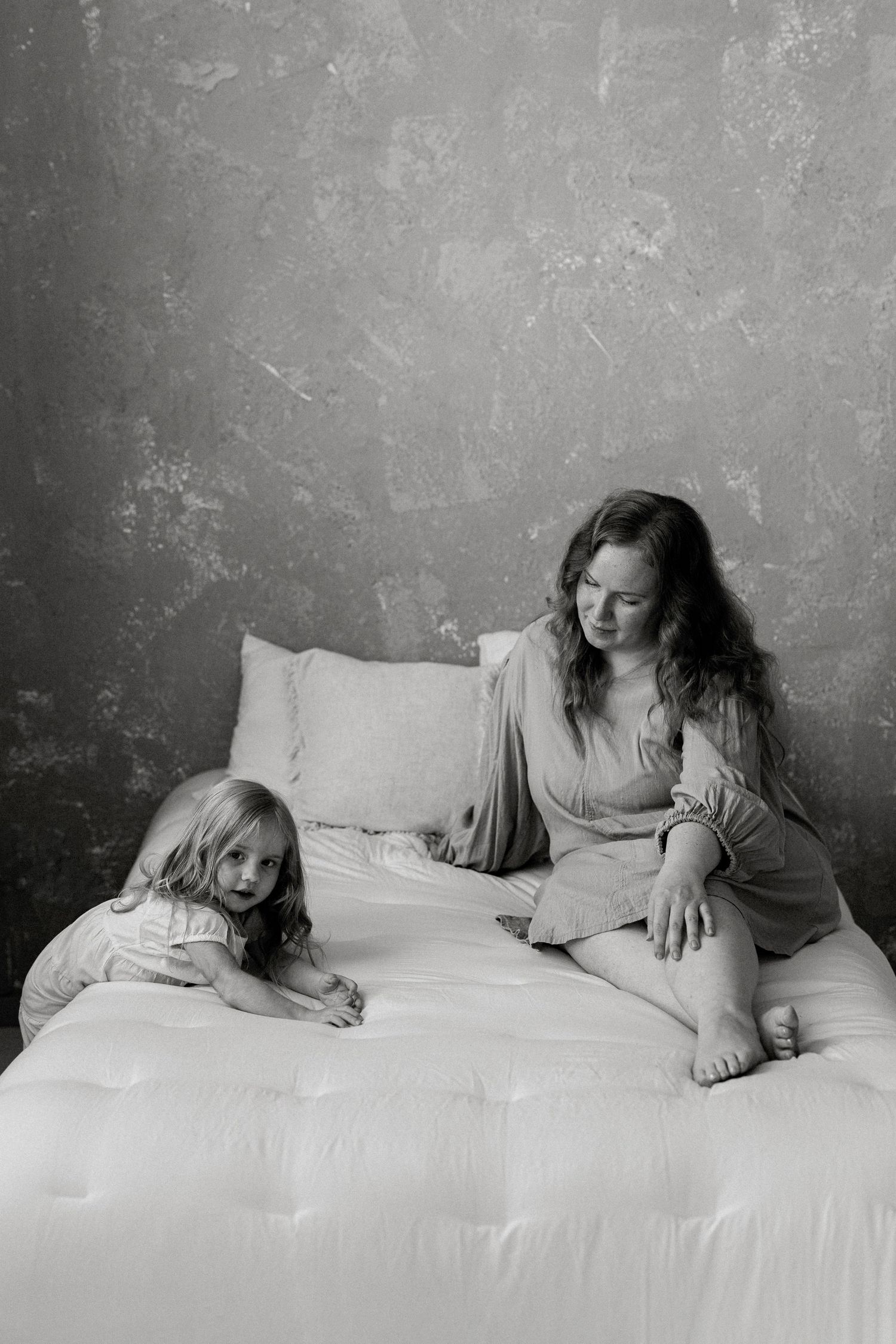 Black and white photo of a mother and young daughter sitting together on a white bed against a textured gray wall.