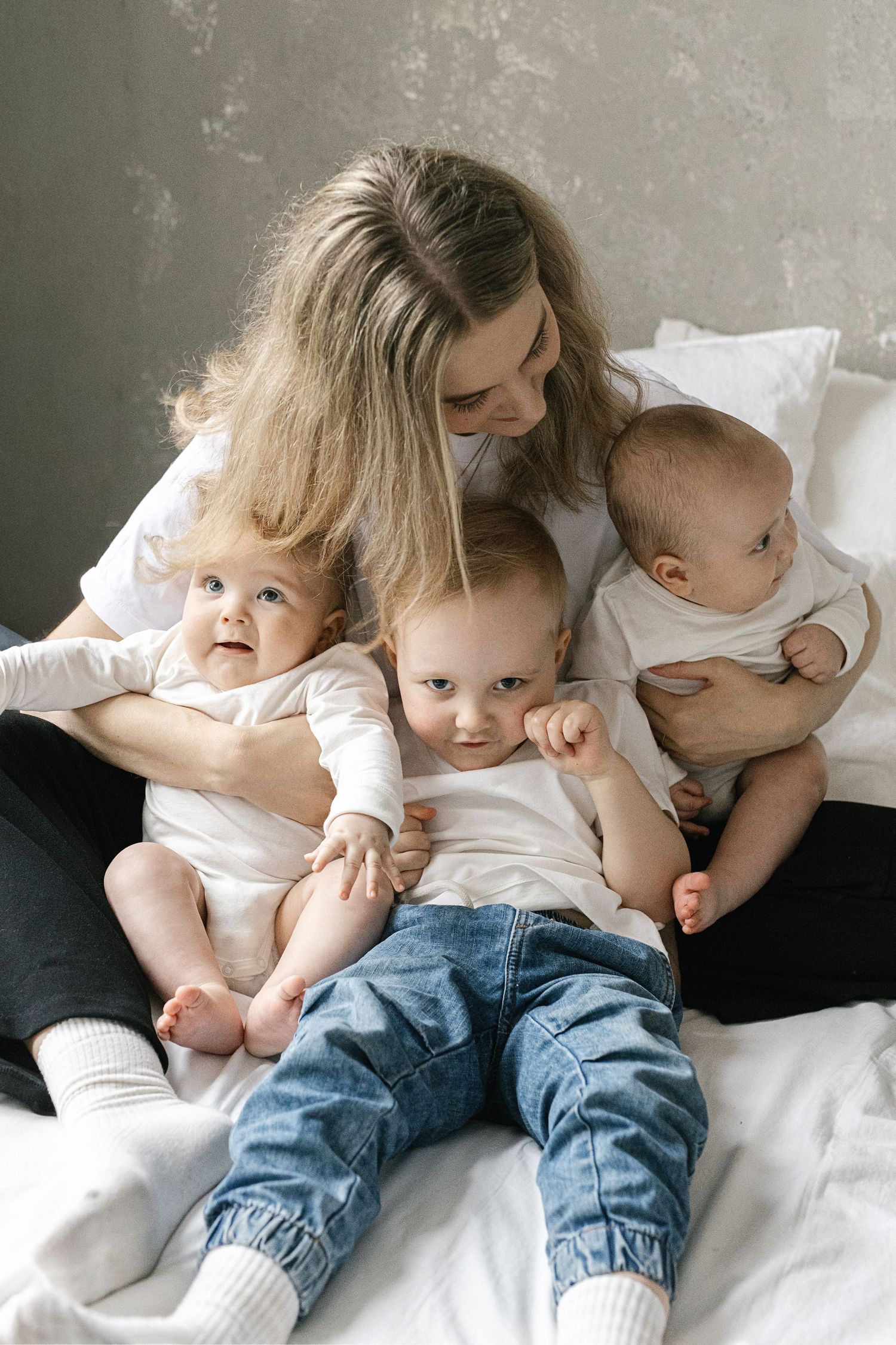 A mother cuddling with three small children on white bedding.