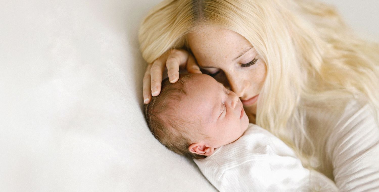 A loving blonde mother gently kisses her sleeping newborn baby while lying on a soft white surface.