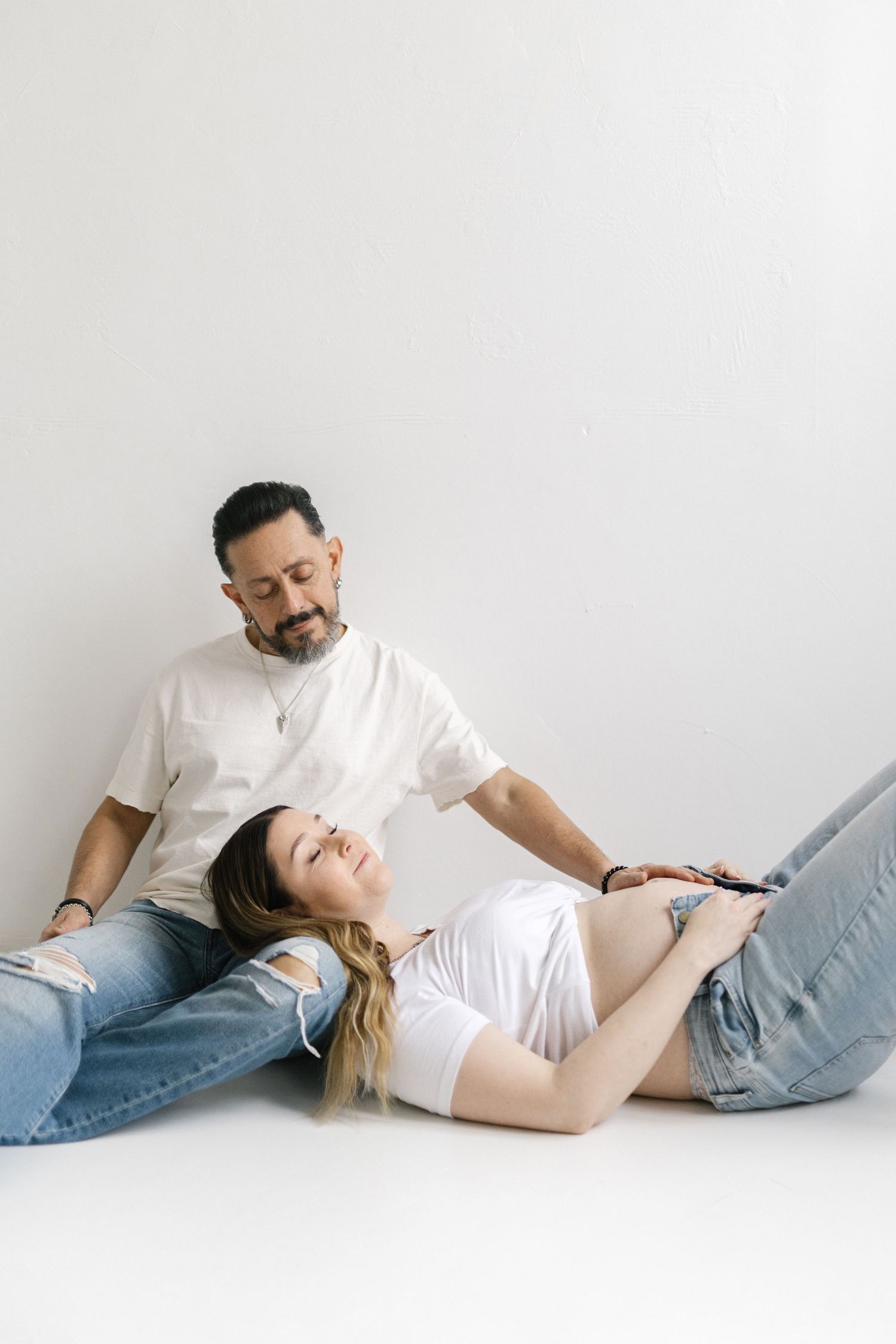 A pregnant woman reclines on her partner's lap on a white floor, both wearing white tees and jeans in a maternity photo.