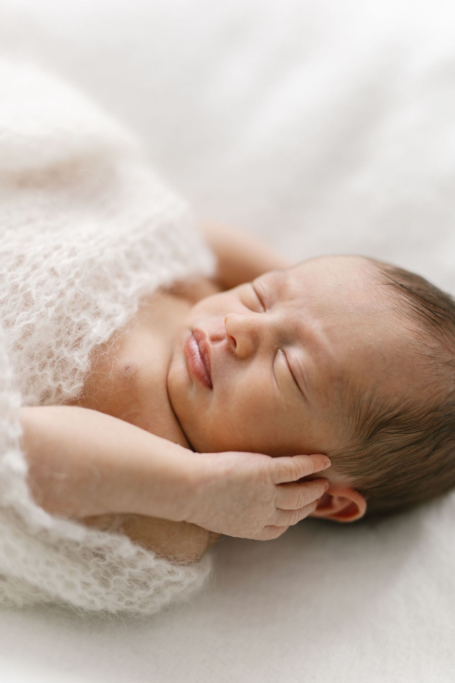 Peaceful newborn baby sleeping wrapped in a white knit blanket with tiny hand resting near face.