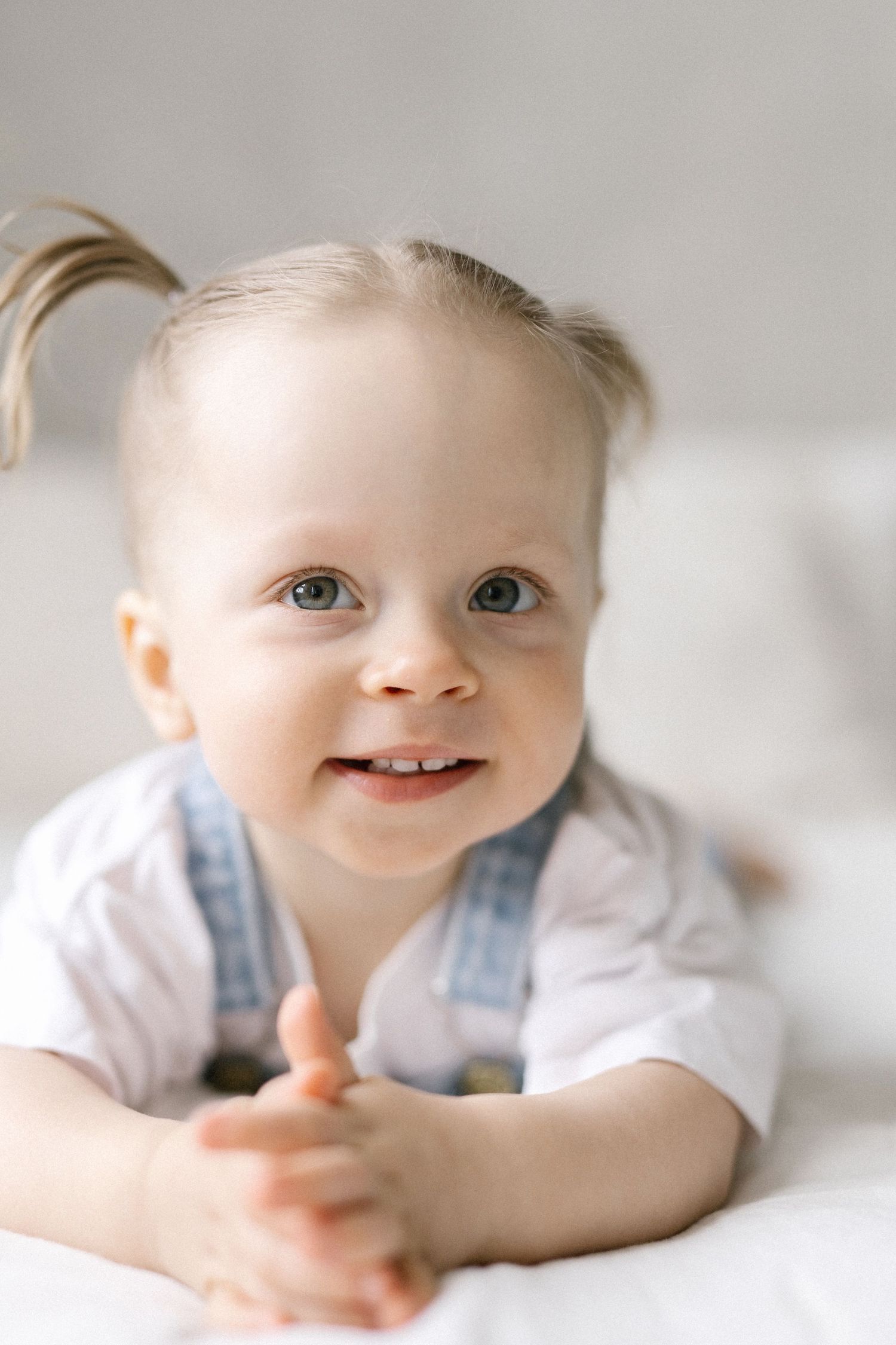 Smiling baby girl with pigtails lying on white surface, wearing denim overalls and white shirt.